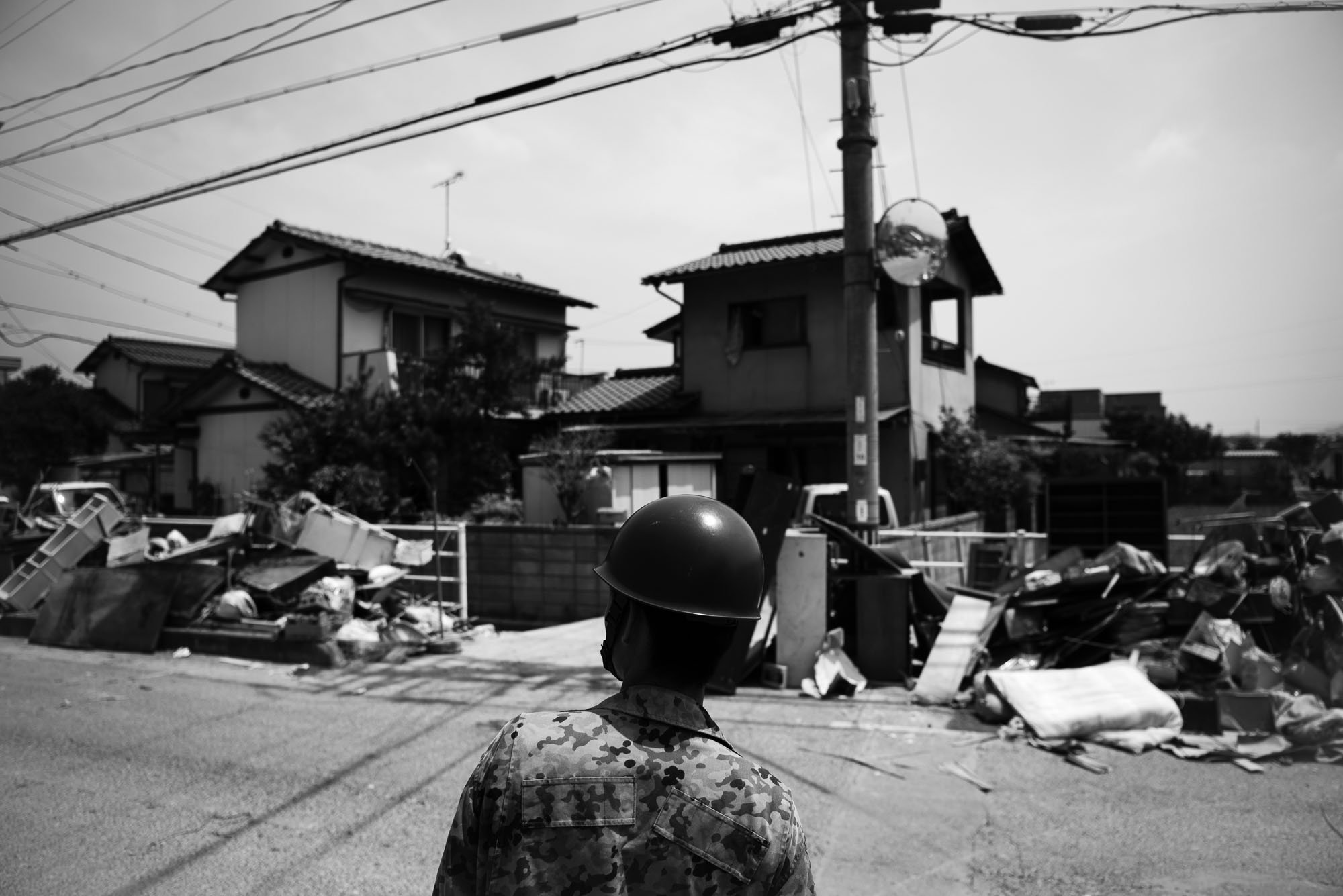 A person in military camouflage uniform and helmet standing on a street, facing a pile of debris and damaged furniture, with houses and power lines in the background.