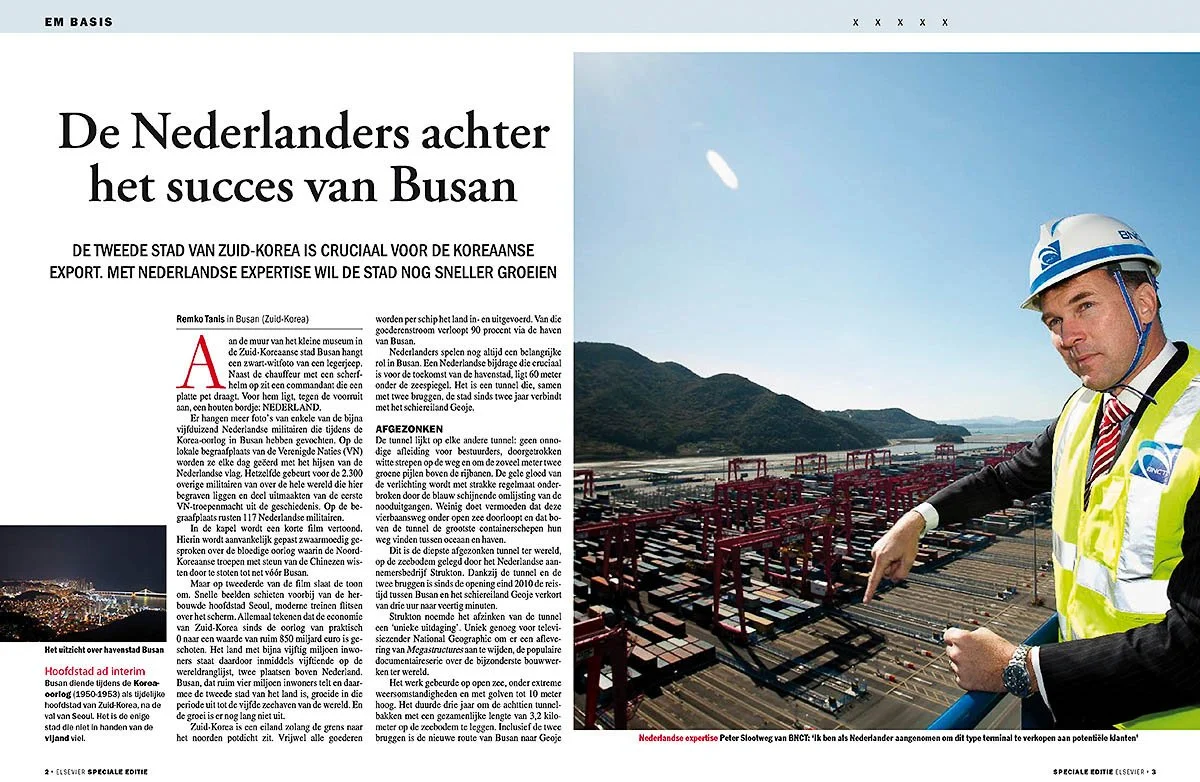 A man wearing a white and blue construction helmet and a yellow safety vest points to a large shipping container yard at a port with mountains and sky in the background.