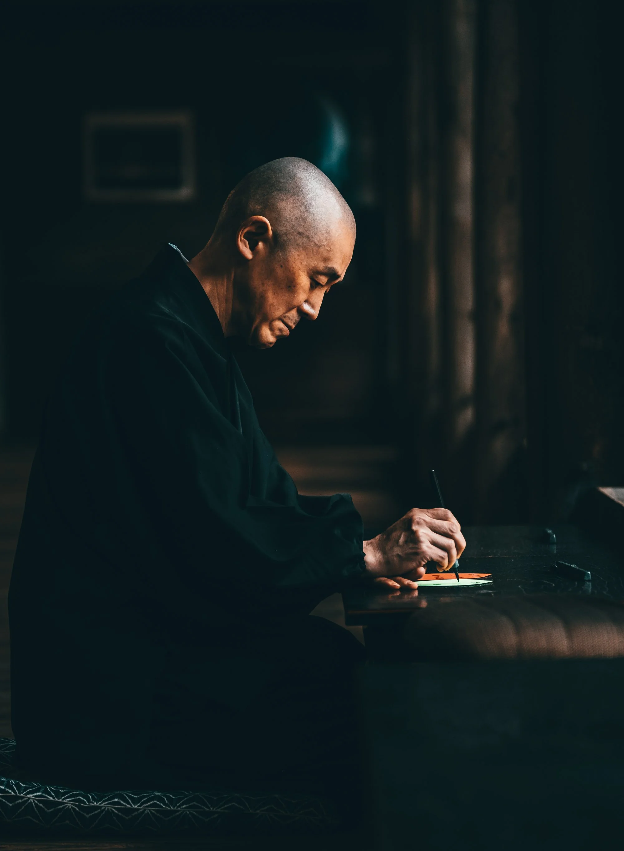 Side-profile portrait of Buddhist monk in dark robes practicing Japanese calligraphy.