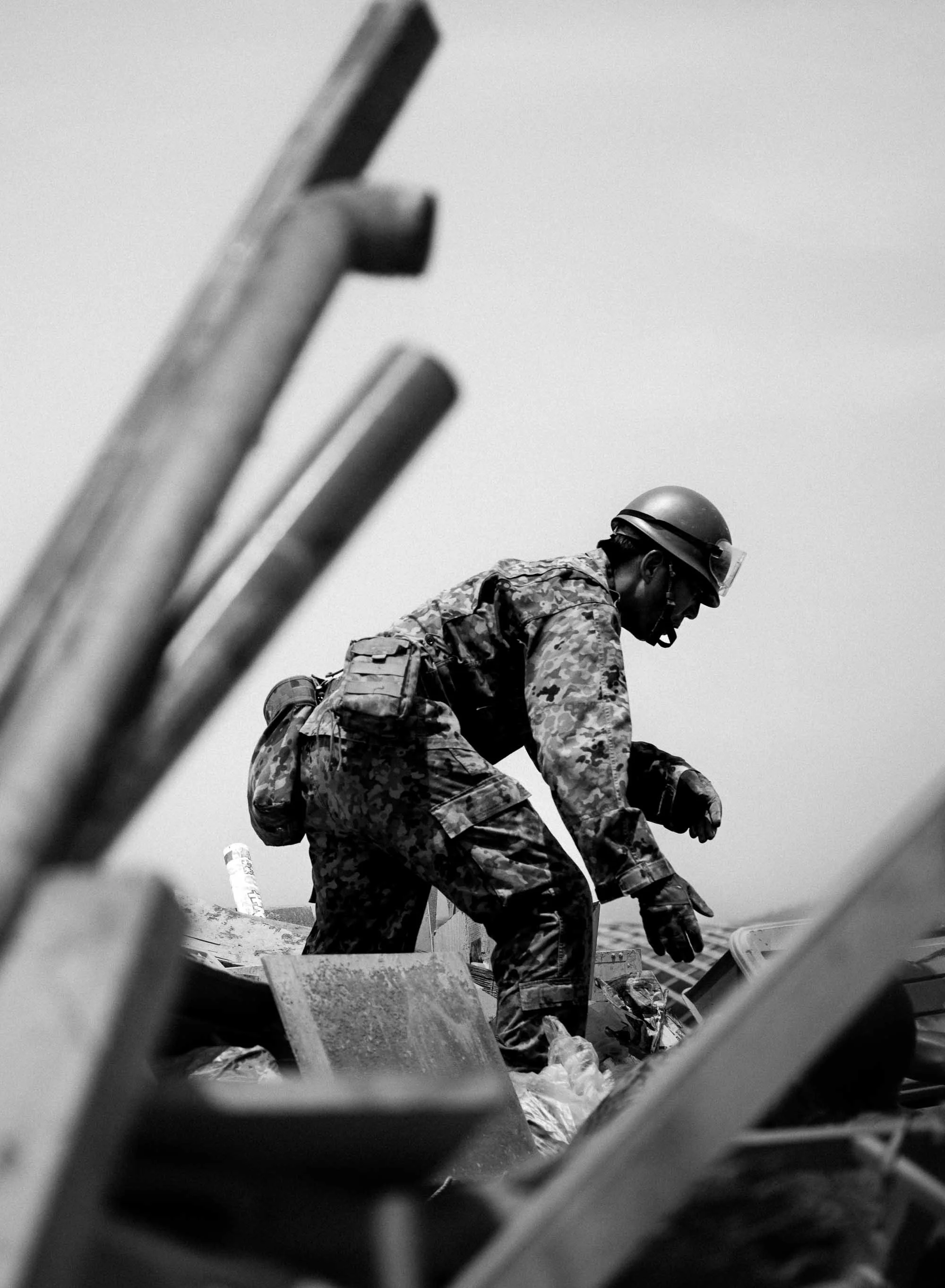 A soldier in camouflage uniform and helmet crouches amid debris and waste in a flood disaster zone.