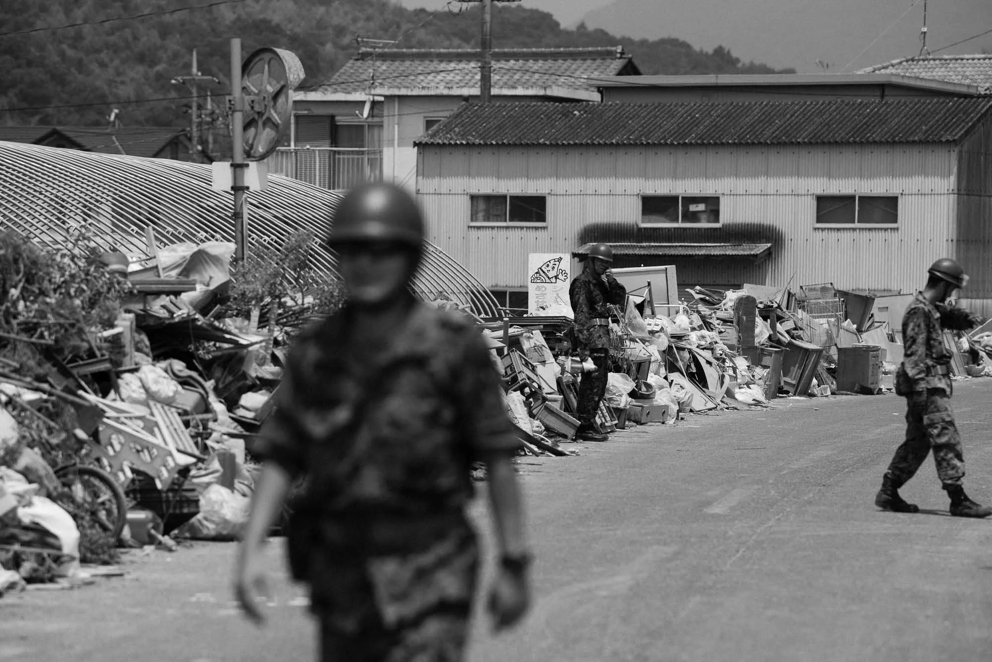 Three soldiers in camouflage uniforms and helmets patrolling a street with a large pile of trash and debris, some buildings in the background, in black and white.