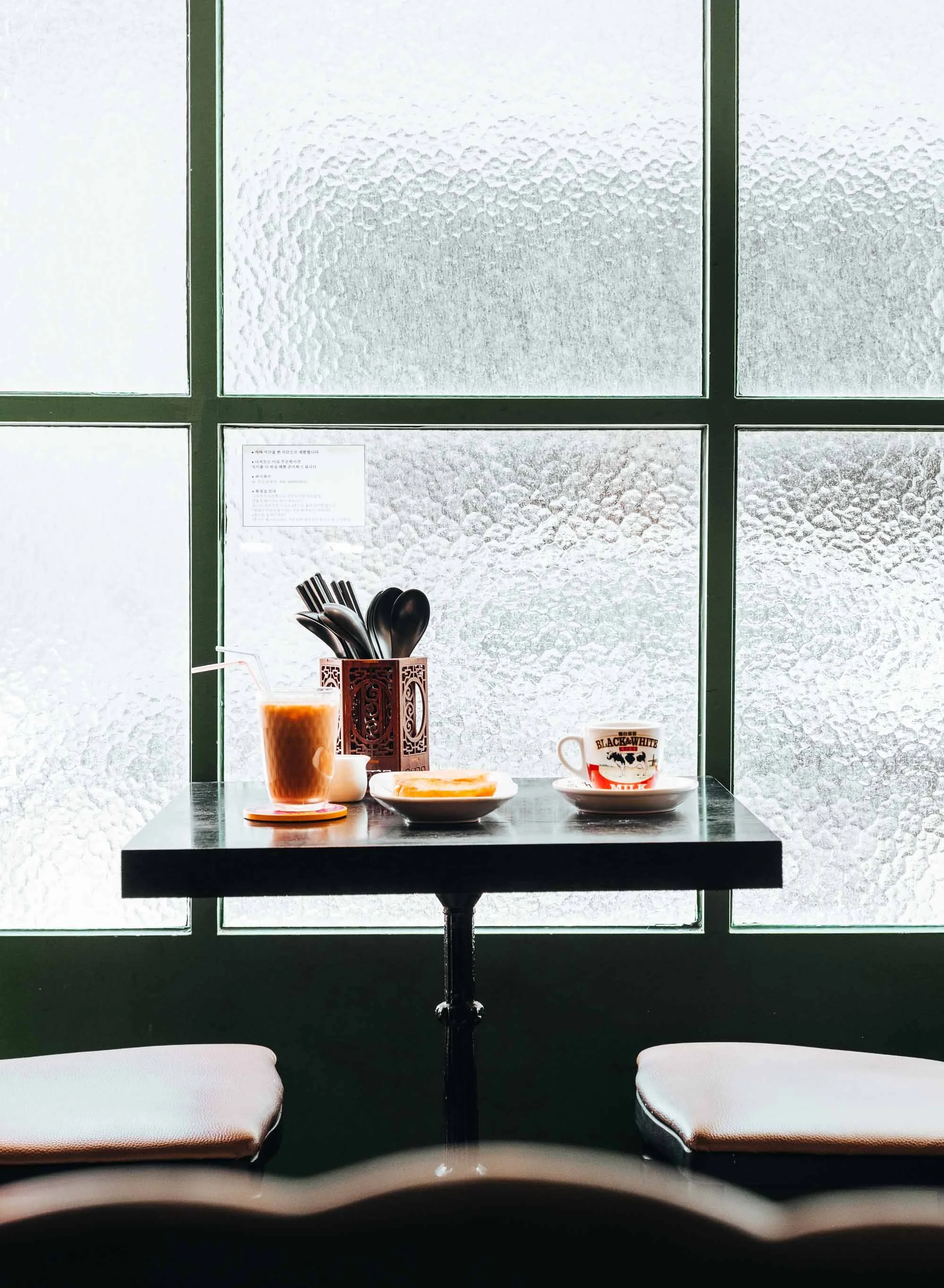 A small black table with a drink, a bowl, and a cup, set against textured glass window panels in a cafe.