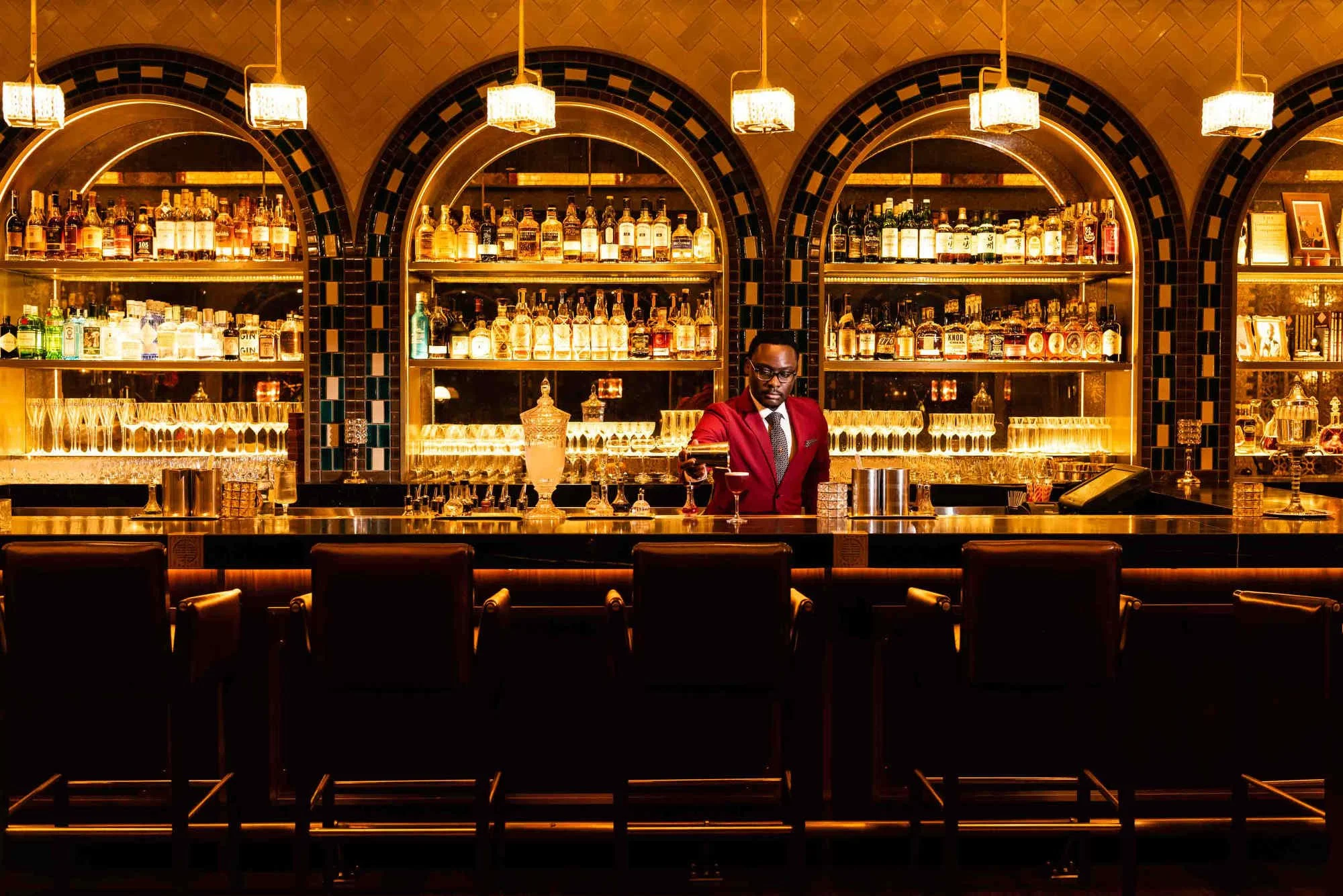 A bartender in a red blazer is preparing drinks behind a luxurious gold-lit bar with shelves of liquor bottles in the background.