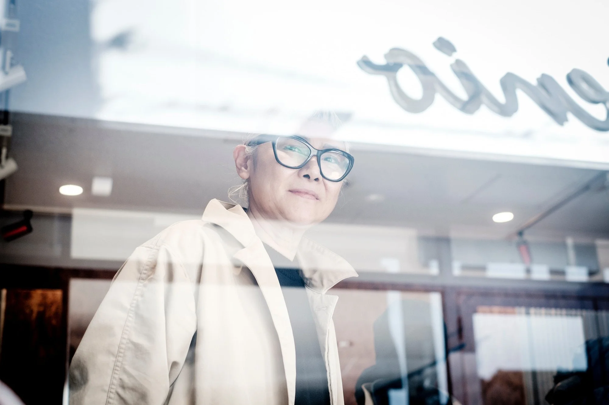A shop owner wearing glasses and a beige jacket looking through a store window.