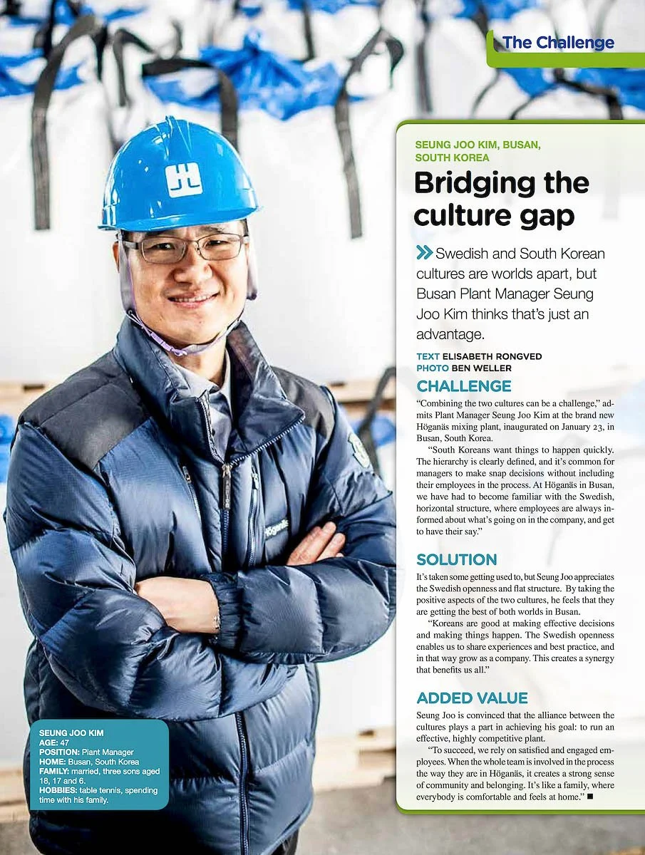A man wearing a blue safety helmet and a blue jacket, standing with arms crossed inside a factory or warehouse with industrial equipment in the background, photographed by Japan industrial photographer Ben Weller.