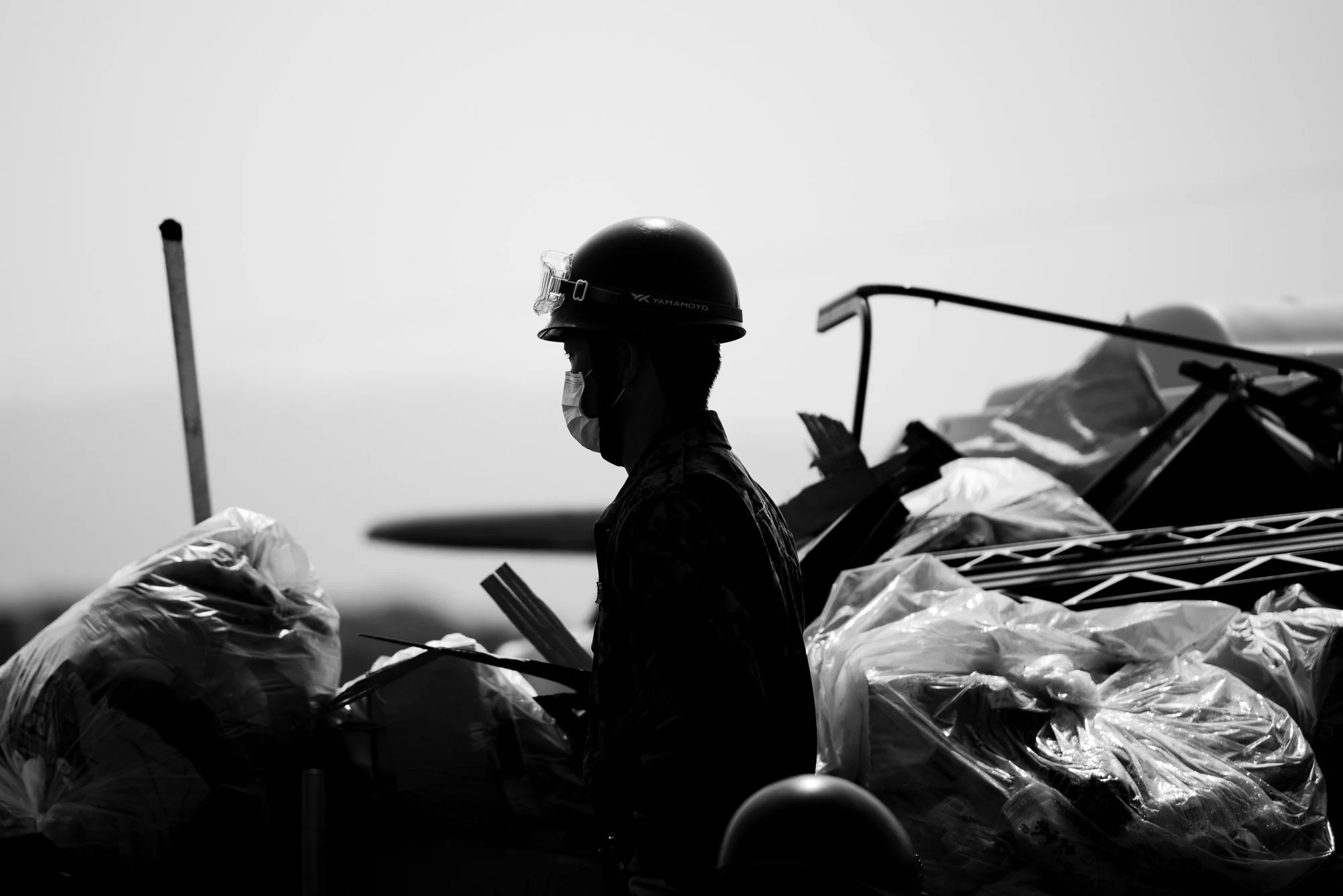 A soldier wearing a helmet and a face mask, standing among bags and equipment, during flood clean up operations in Okayama, Japan.