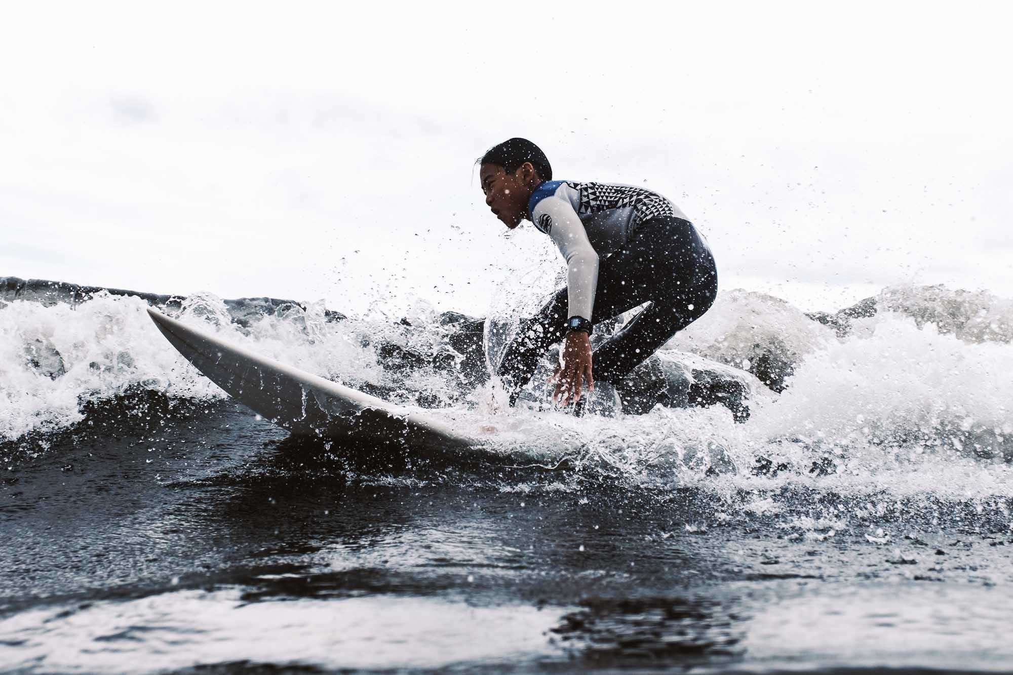 A young boy surfing on a wave in the water outside under a cloudy sky by Japan-based sports photographer Ben Weller.