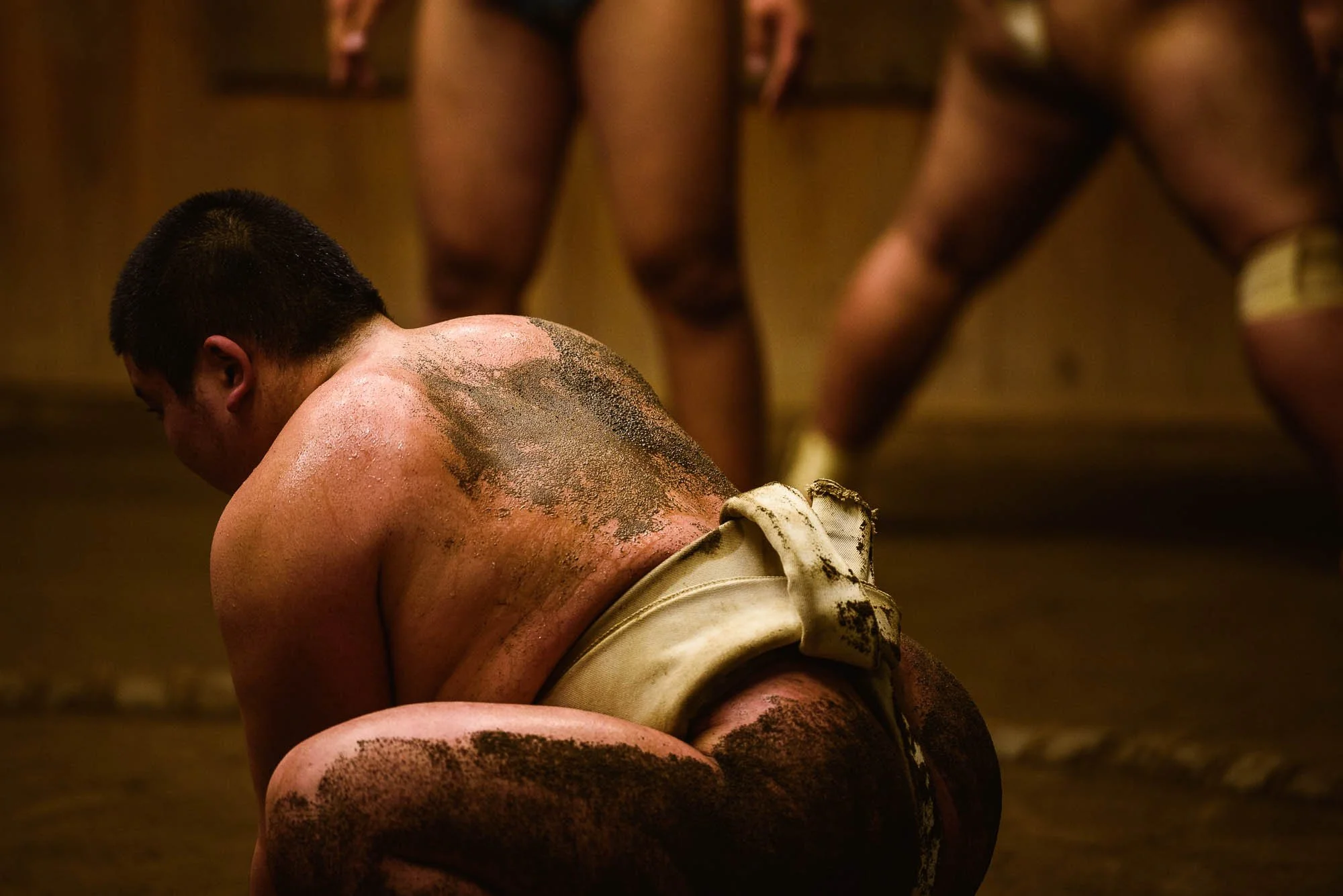 Young man squatting in a traditional Japanese sumo wrestling ring, with visible black dirt or clay on his body, wearing a white mawashi, preparing for a sumo match, with other wrestlers in the background.