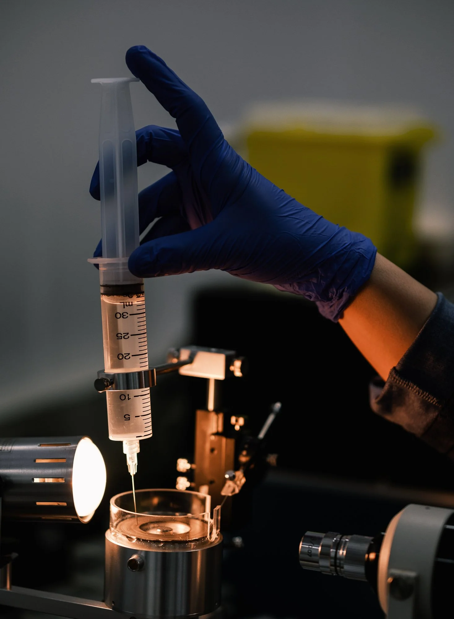 A scientist's hand wearing a blue glove holding a syringe with a pink liquid, positioned over a petri dish on a lab table, with laboratory equipment visible in the background.