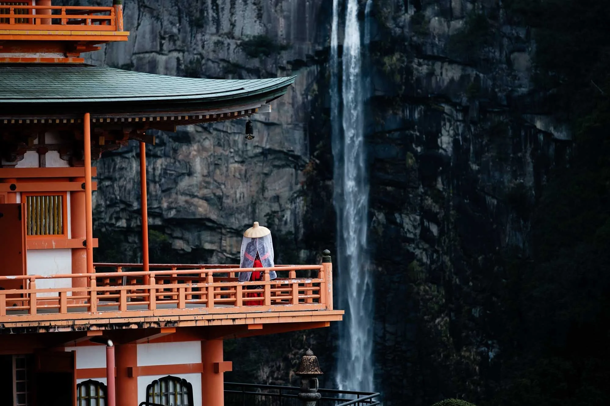 A woman wearing a traditional Japanese hat and veil stands on the balcony of a red pagoda facing a towering waterfall in the background.