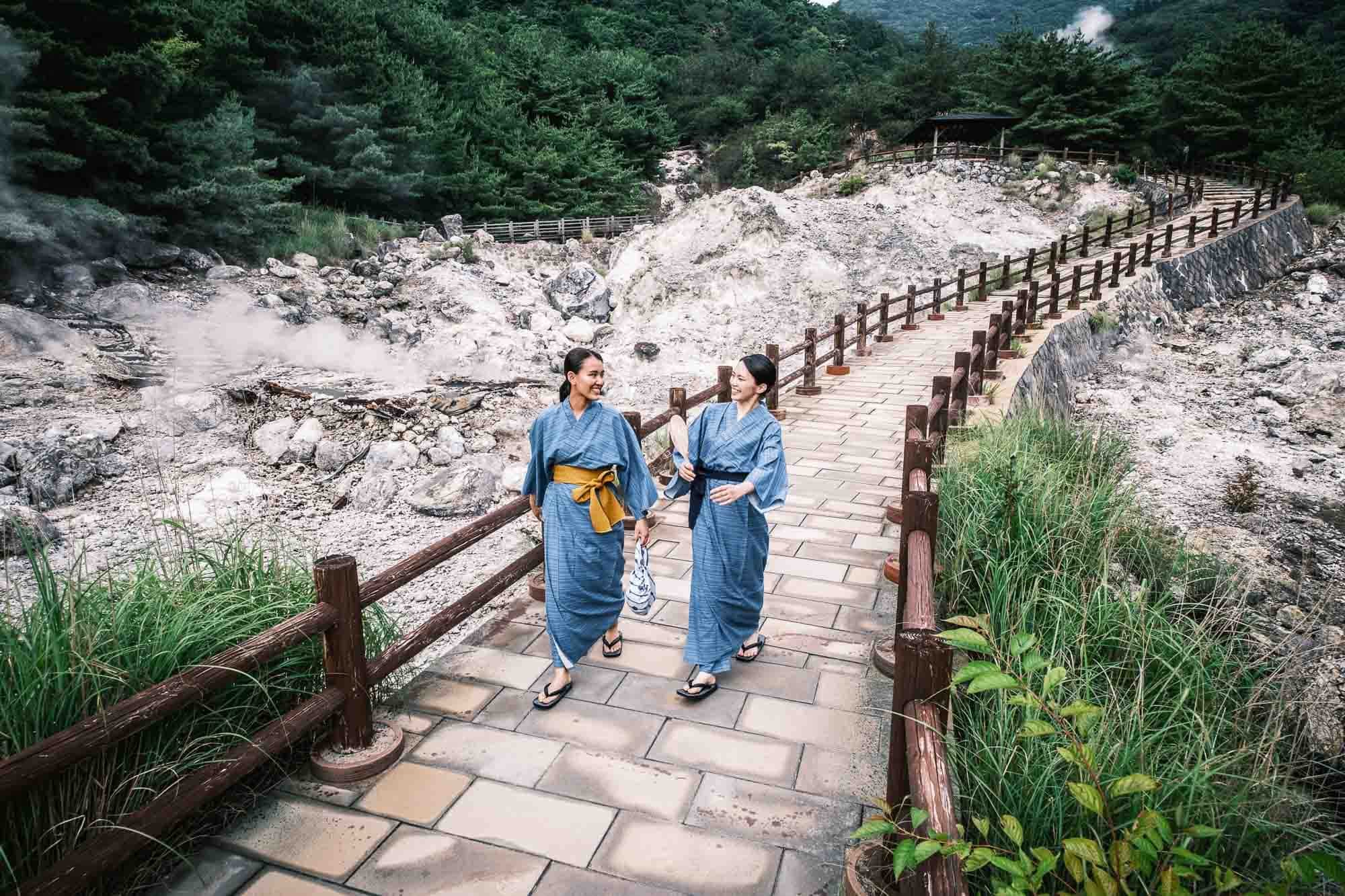 Two women wearing traditional Japanese yukata clothing walk on a path  between natural hot springs in Unzen, Nagasaki.