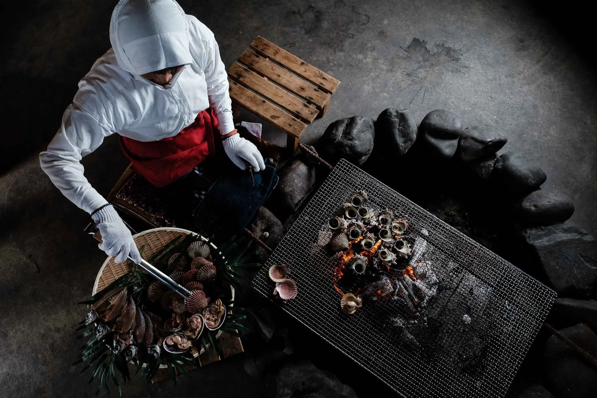 Ama diver woman in traditional diving attire grills shellfish over a stone hearth in Iseshima, Japan.
