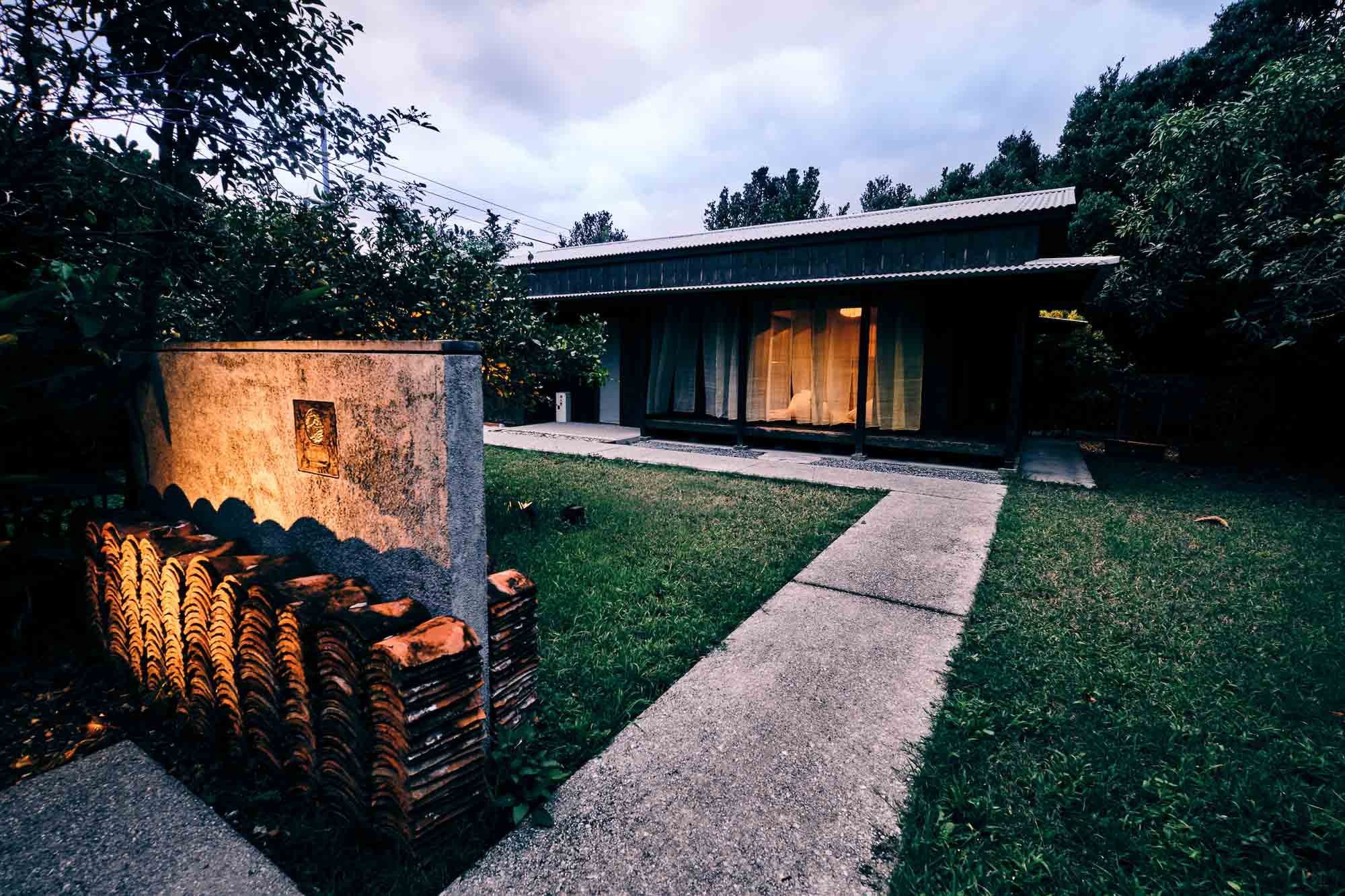 Traditional Okinawan kominka home that has been converted into a boutique hotel. The yard features a concrete walkway and a stone wall with clay roof tiles stacked at the base, under a cloudy evening sky.