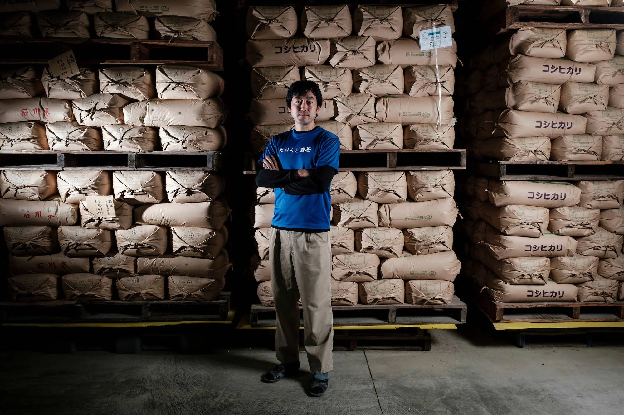 Full-body editorial portrait of a Japanese man standing inside a rice storage facility.