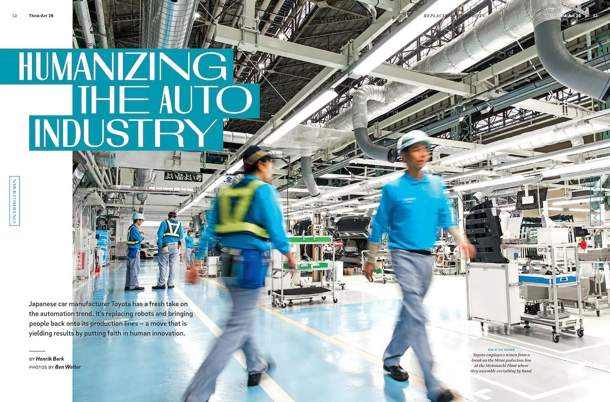 Inside a Toyota car manufacturing plant, workers in blue uniforms and safety helmets walk through an industrial space with machinery, car parts, and a ceiling filled with pipes and ducts.