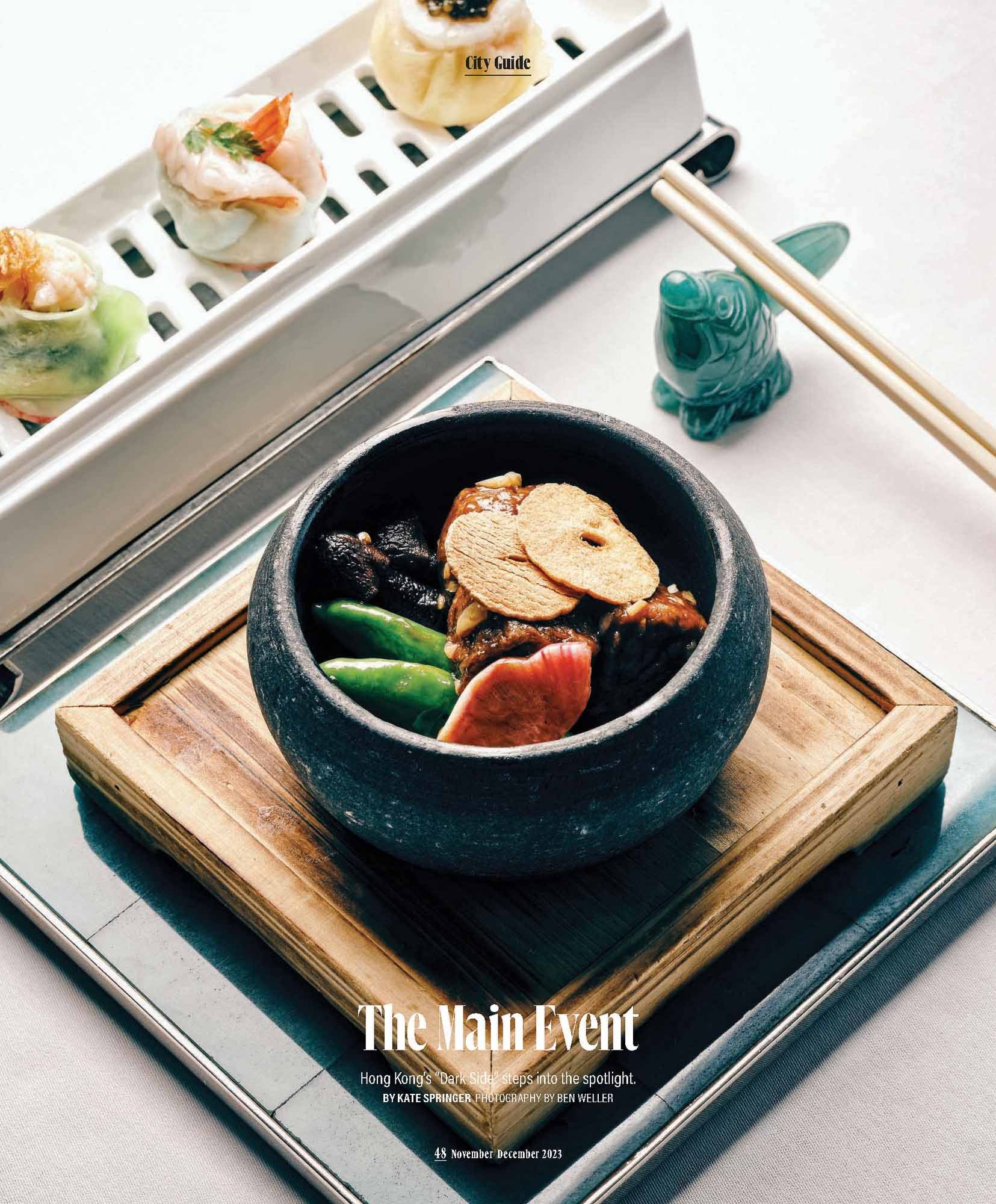 The lead image for a magazine story on Hong Kong, a black stone bowl filled with Asian cuisine, placed on a wooden tray, with vegetables and sliced mushrooms, surrounded by a white table setting and chopsticks.