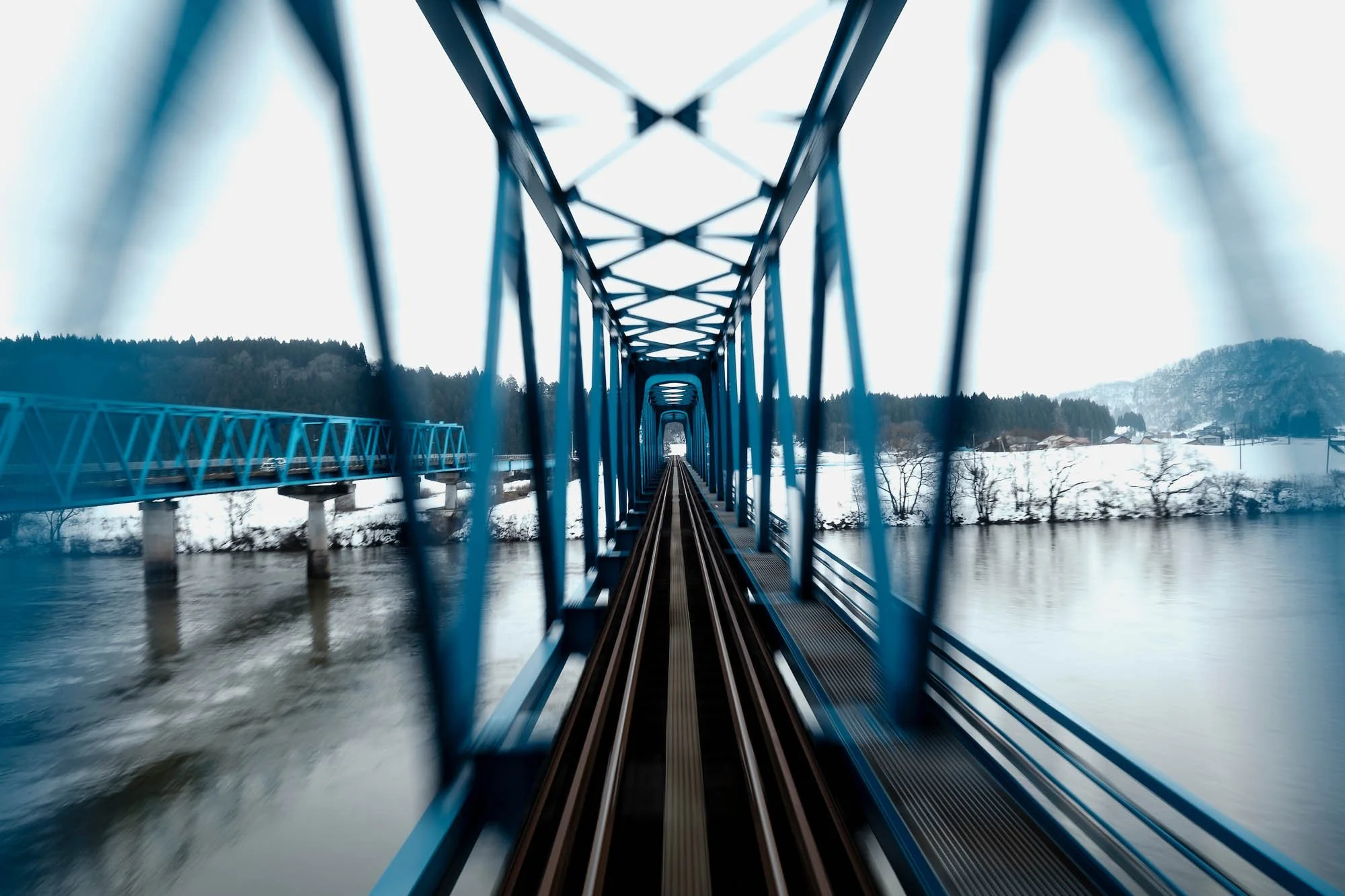Point of view shot from a train traveling at high speed across a railroad bridge crossing water in a snowy winter landscape.