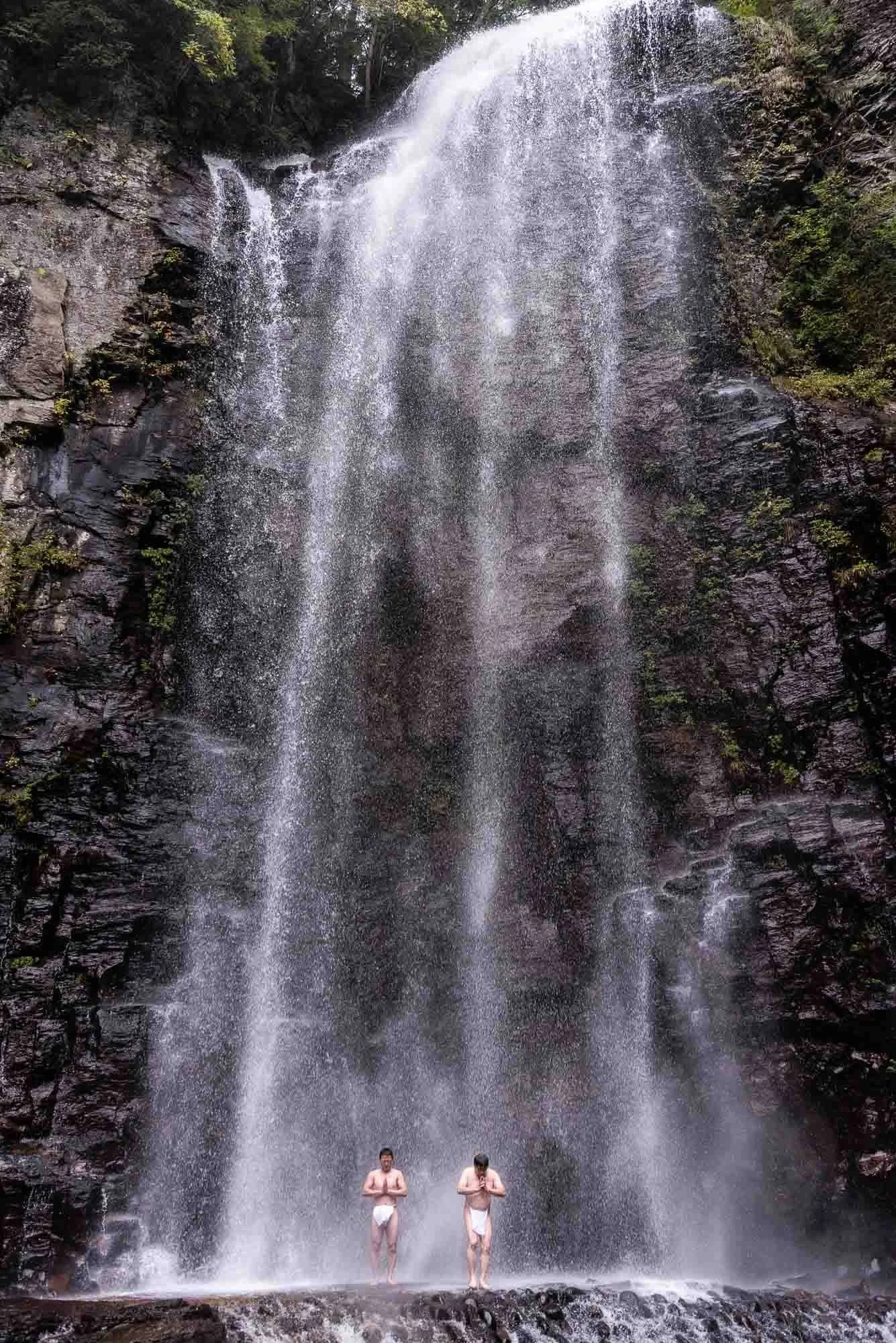 Two men in loincloths stand in prayer under a plummeting waterfall. Shot on assignment by Japan travel photographer Ben Weller.