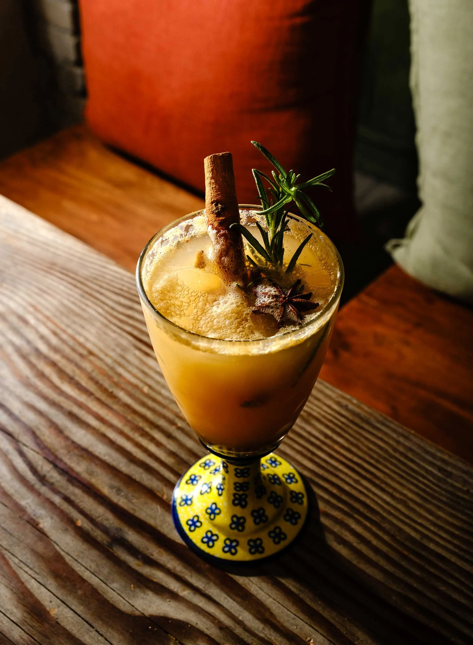 A creamy yellow cocktail garnished with a cinnamon stick, star anise, and a sprig of rosemary, served in a decorative glass on a wooden table.