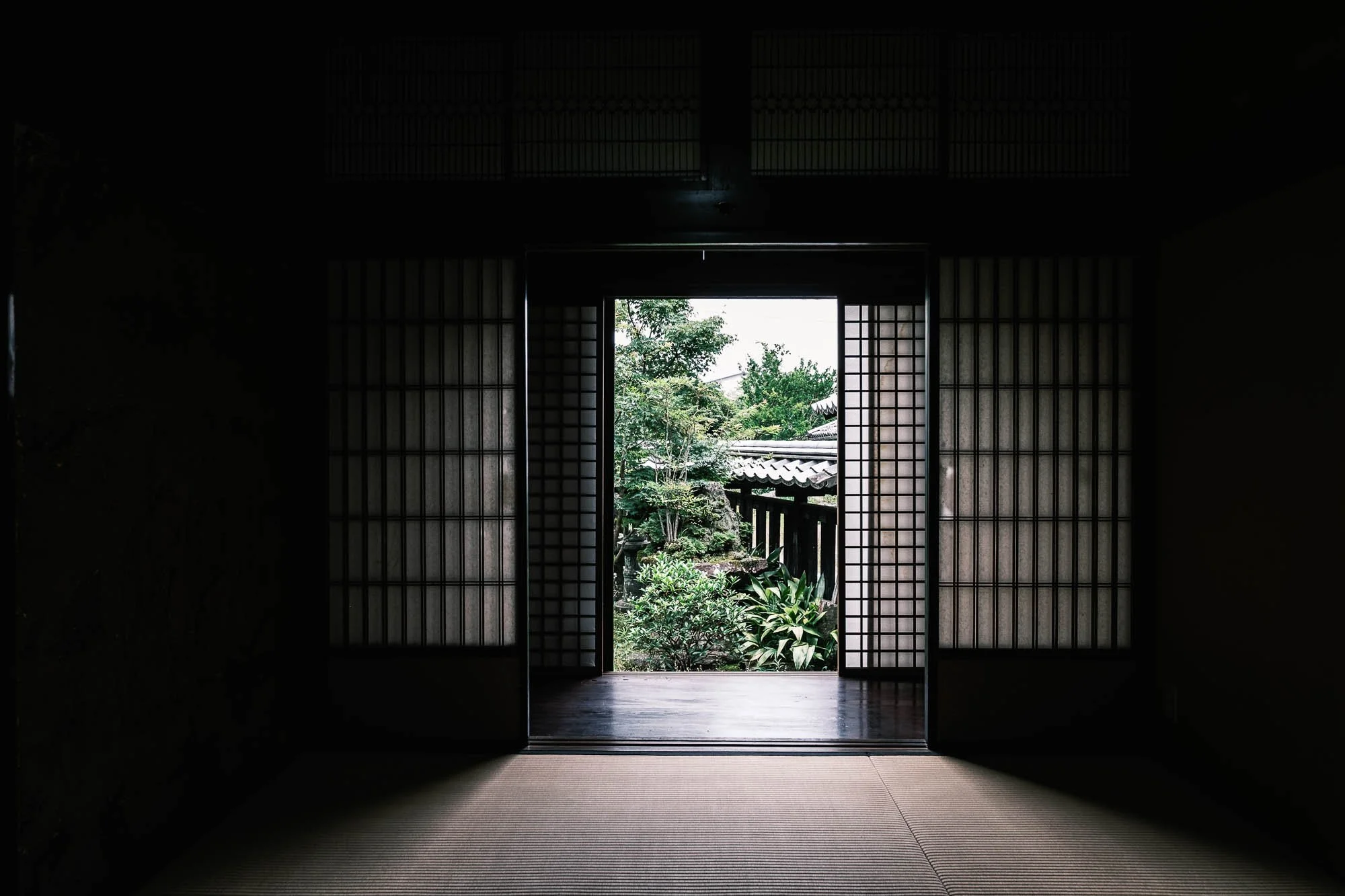 Interior view of a traditional Japanese room with shoji doors open to a garden with trees, shrubs, and a pathway.