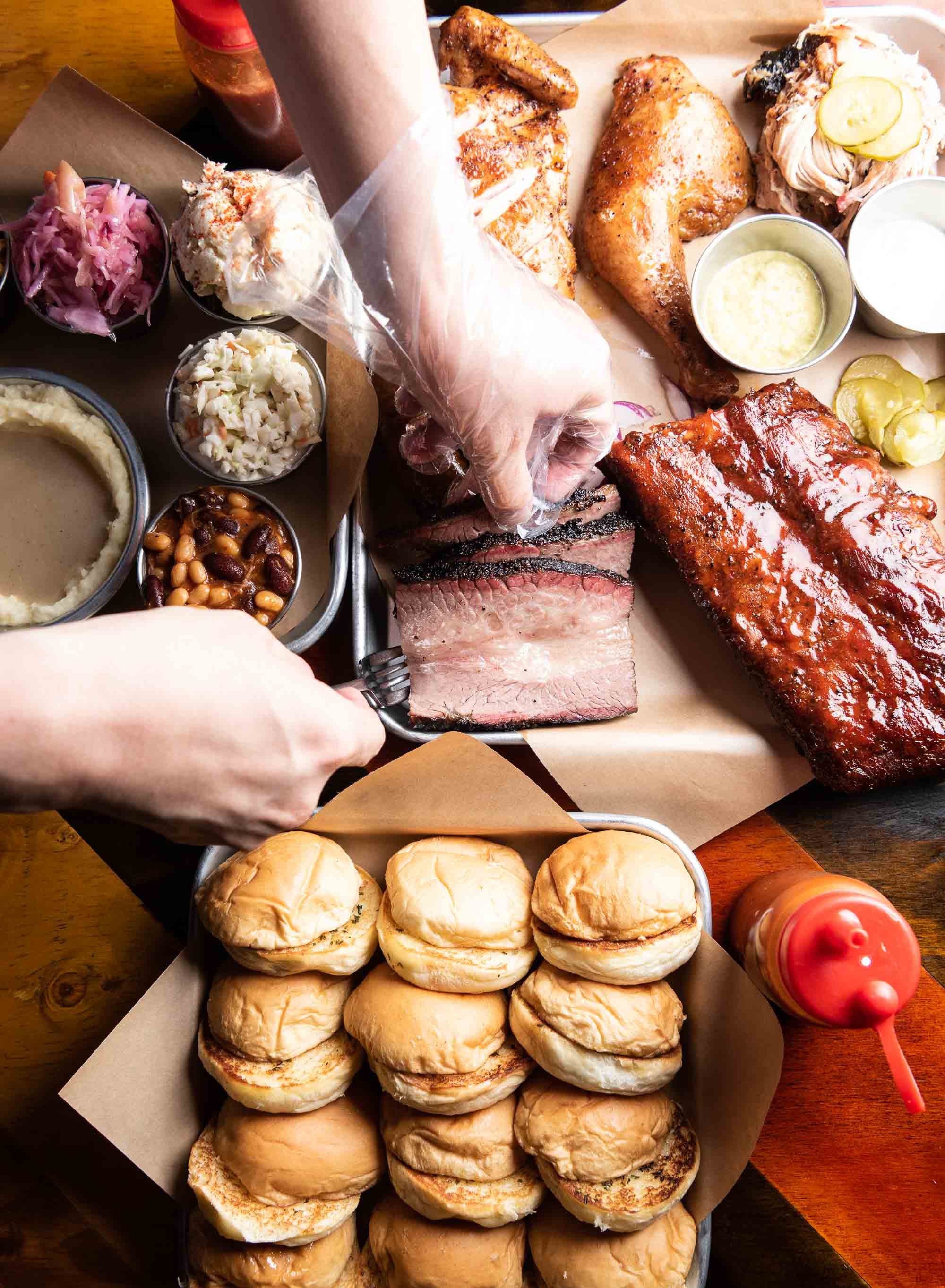 A spread of barbecue food including smoked meats, buns, side dishes, and condiments on a wooden table.