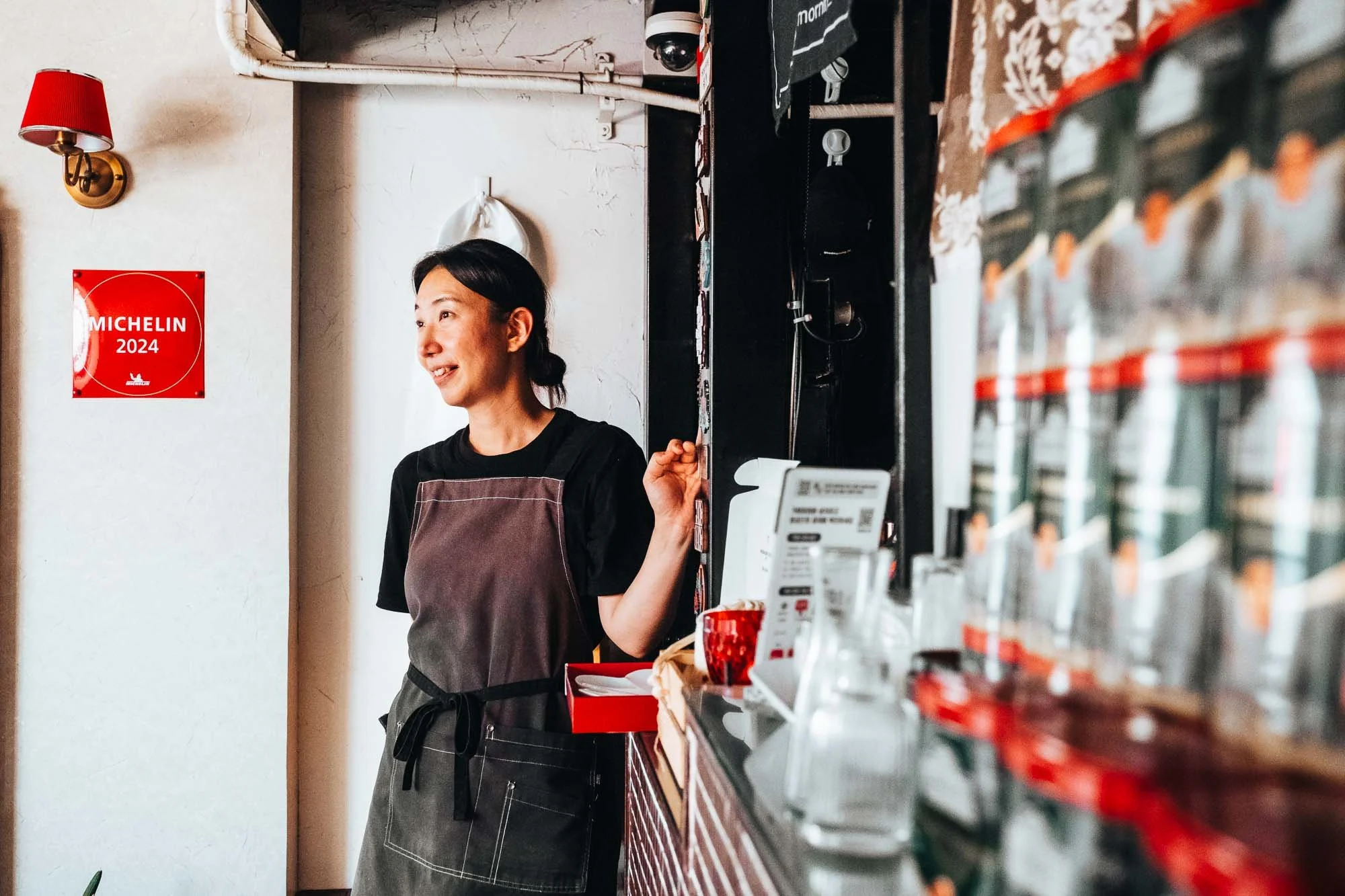 A woman working behind the counter of a restaurant or cafe, wearing a black shirt and gray apron, smiling and talking. There is a red Michelin 2024 sign on the wall behind her, and shelves with various items on her right.