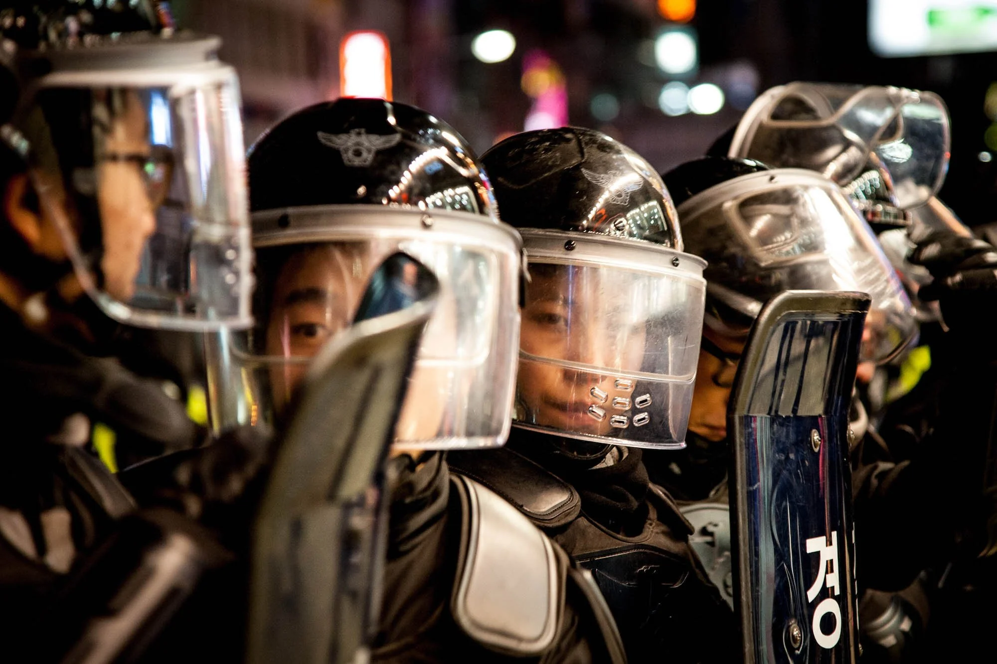 Line of Korean riot police wearing black uniforms and helmets with visors, holding riot shields, standing in a row at night,  photographed by Ben Weller, editorial photographer in Japan.