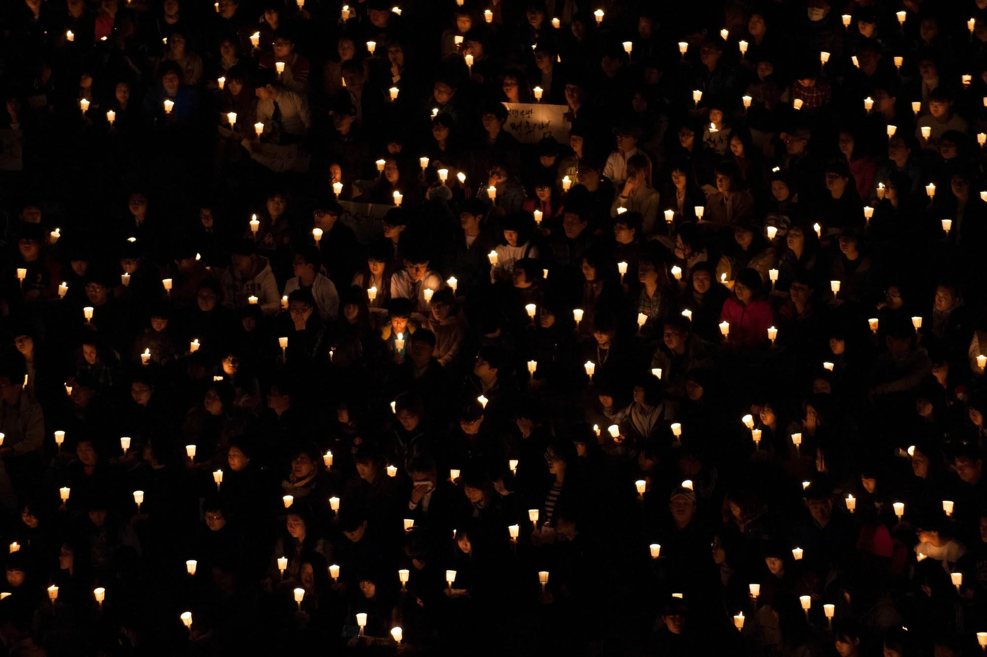 A large group of people gathered in darkness, holding lit candles in a vigil for students killed in the Sewol Ferry Disaster in South Korea.