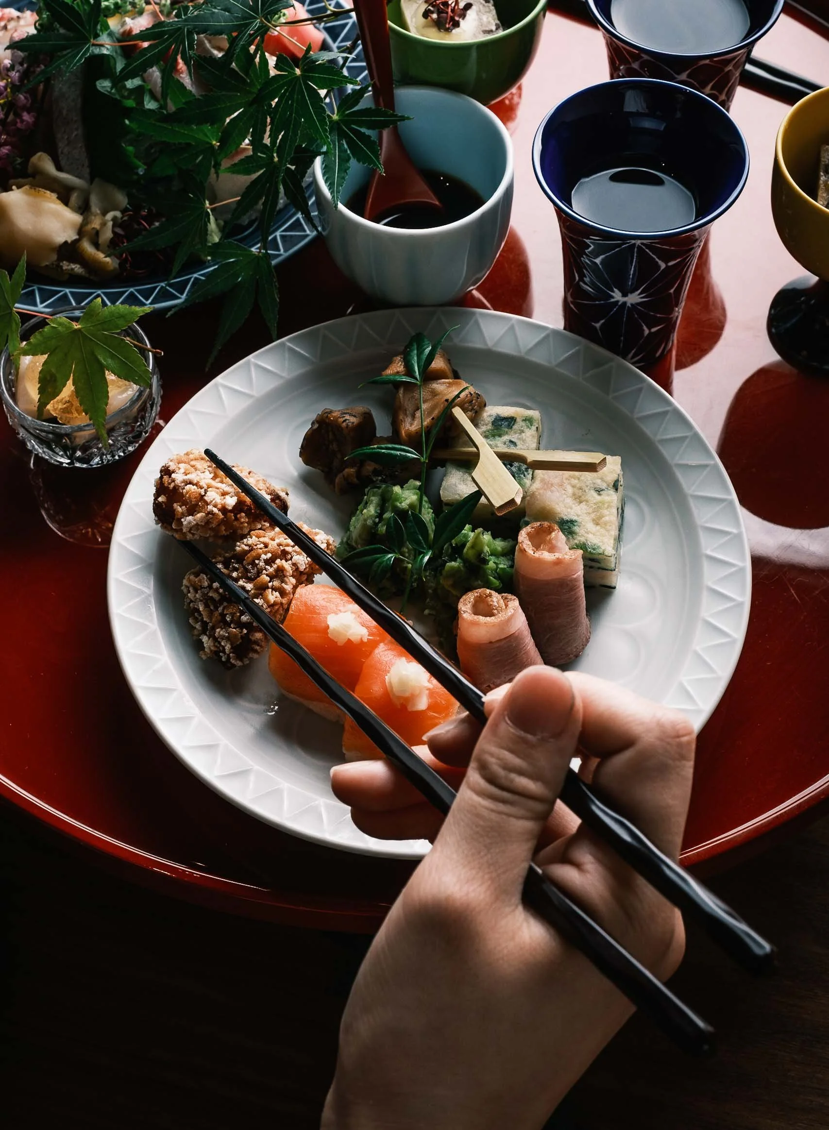 A hand holds black chopsticks over a white plate with assorted Japanese food, including salmon sashimi, rolled tuna, fried chicken, and various garnishes. The table has several cups and bowls with different sauces and drinks, decorated with green lea