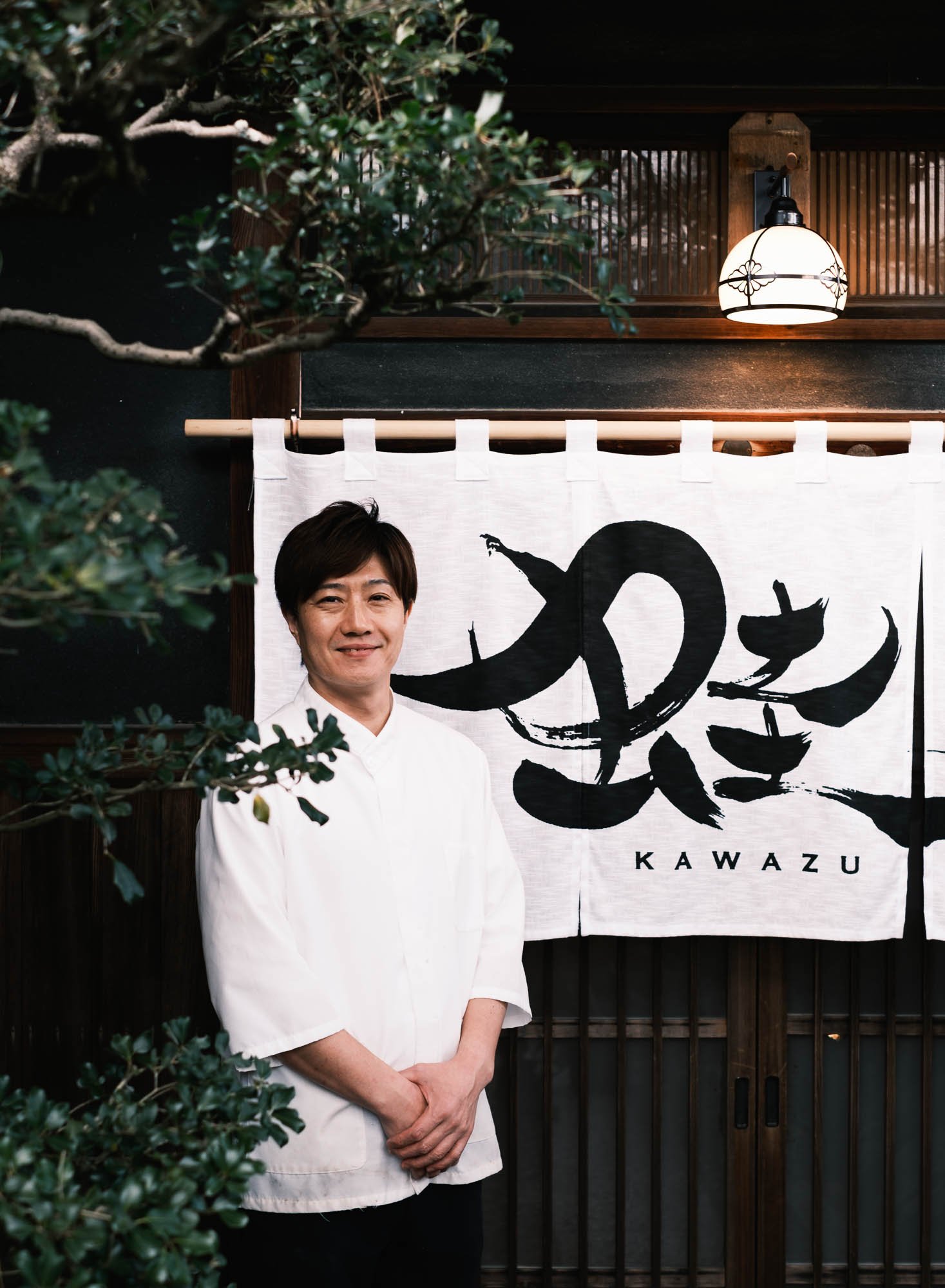 A Japanese chef standing in front of a traditional noren curtain with the word "KAWAZU" in English and Japanese calligraphy. The chef is smiling and wearing a white chef's coat.