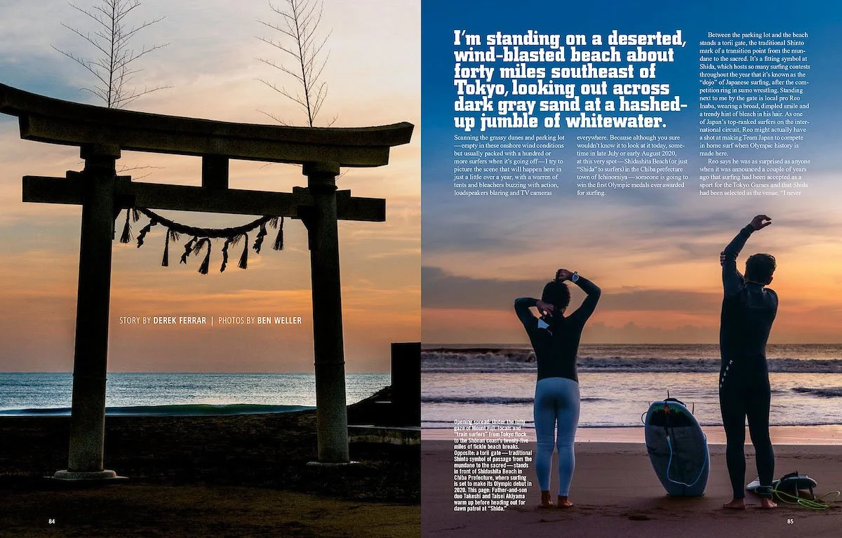 A traditional Japanese torii gate stands on a deserted wind-blown beach at sunset, with two surfers preparing near the shoreline, silhouetted against the sky. Beach and ocean visible in the background.