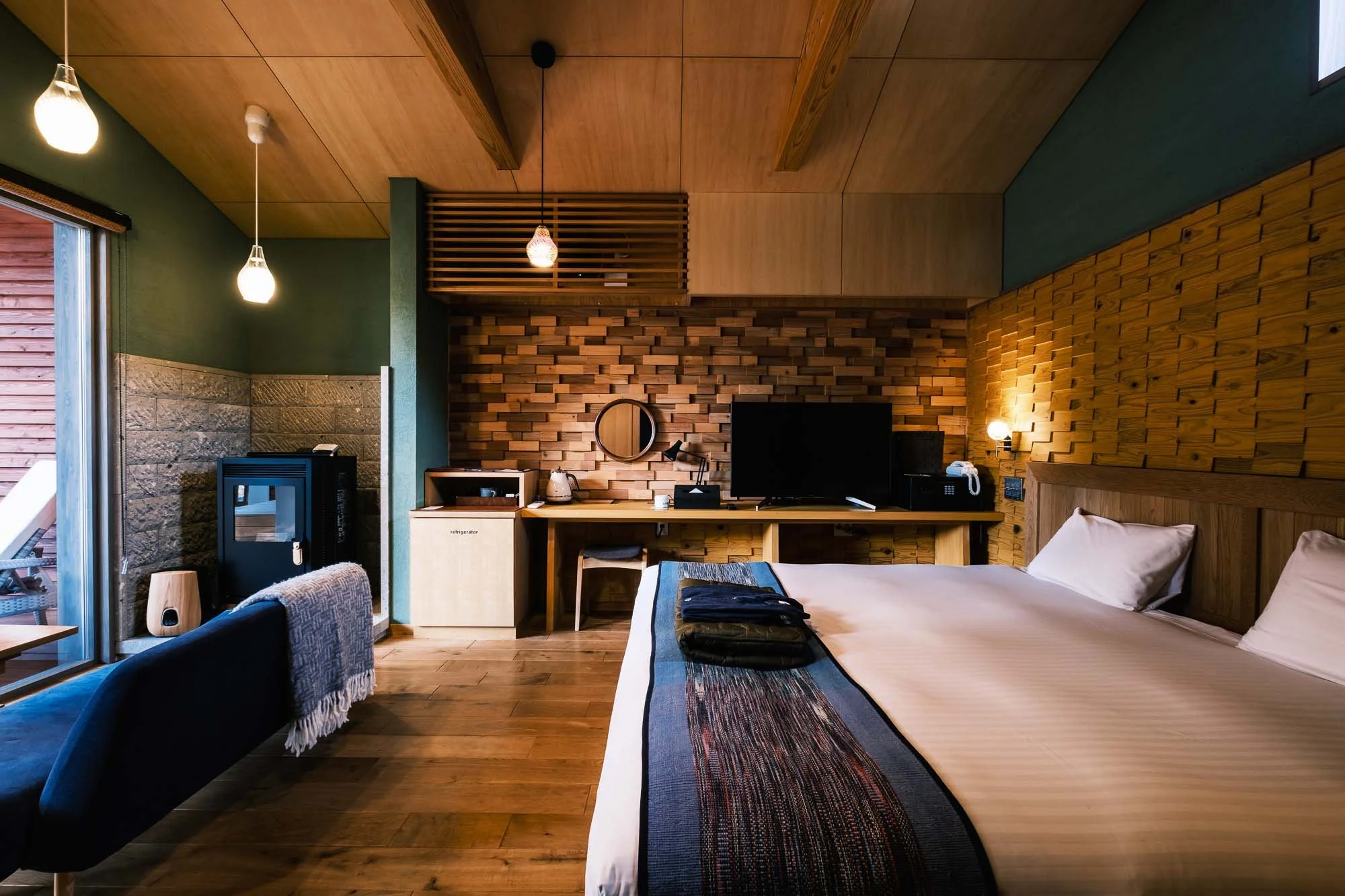 Interior of a cozy hotel room with wooden walls and ceiling, a bed with white linens, folded clothes, a TV on a wooden desk, a small refrigerator, a microwave, a kettle, a sitting area with a blue armchair, and a sliding glass door leading outside.