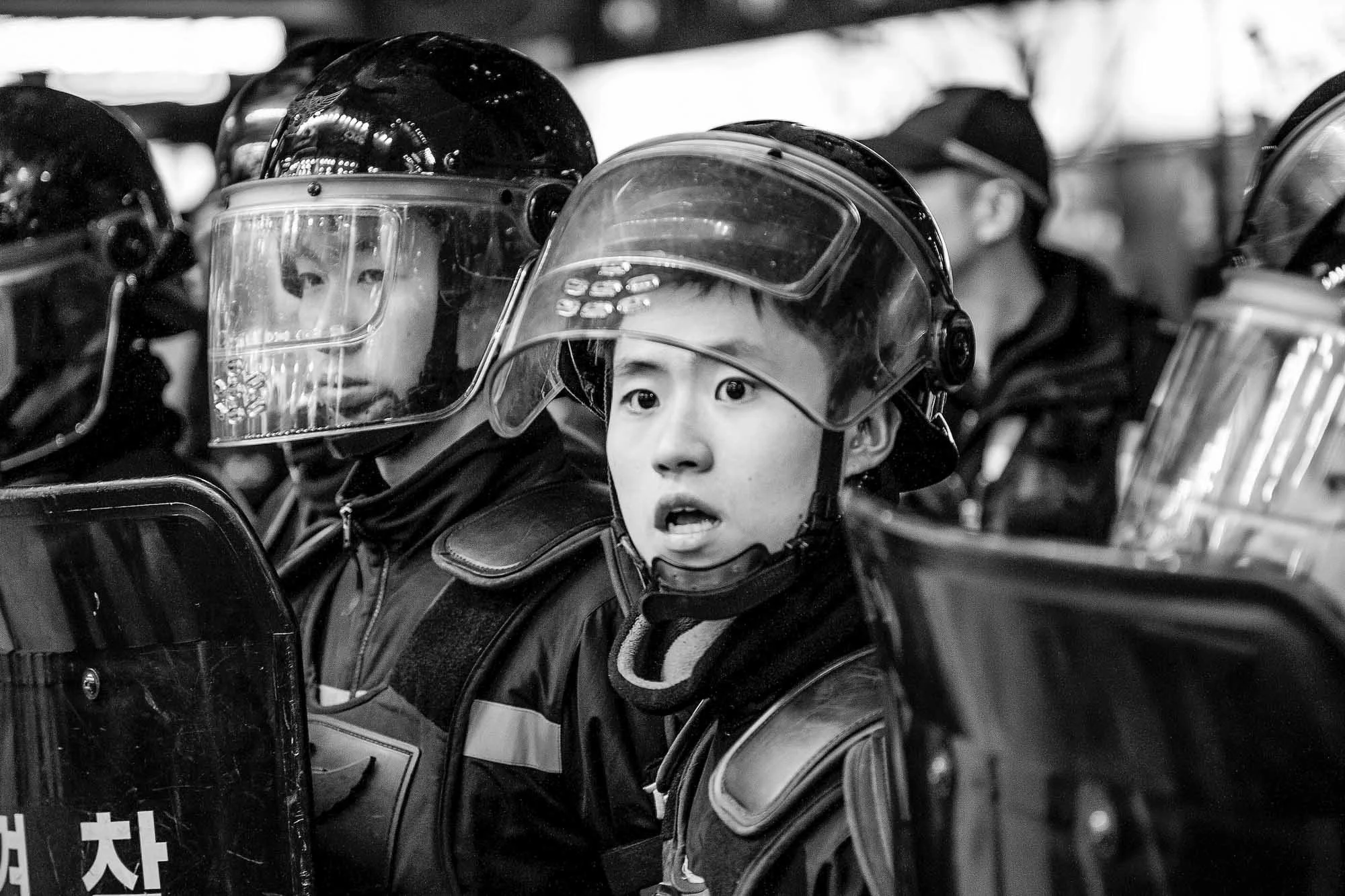 A group of young police officers in riot gear, including helmets with visors, standing closely together in a line.