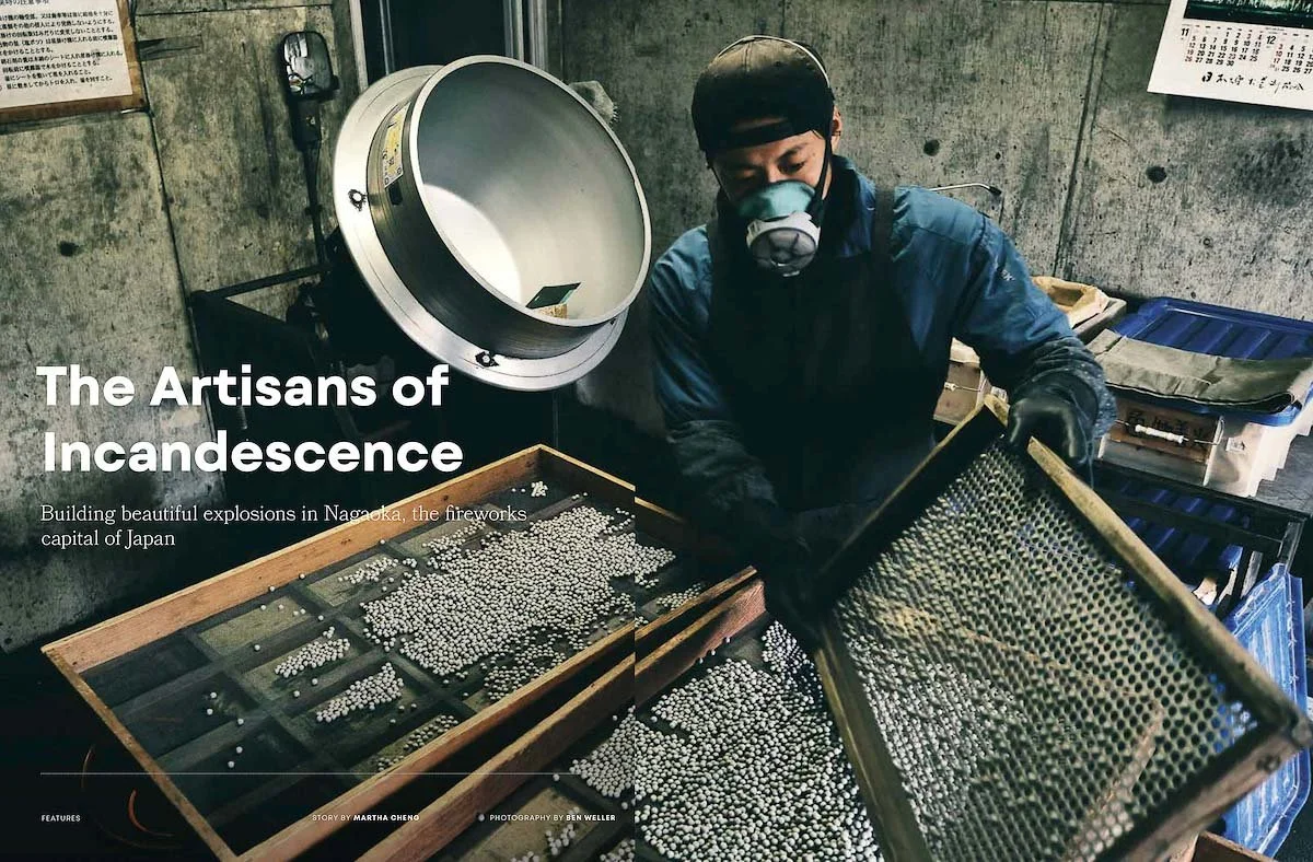A person wearing a protective mask and gloves, working with a tray of small white pellets or fireworks components in a workshop with concrete walls and a large metal tube in the background.