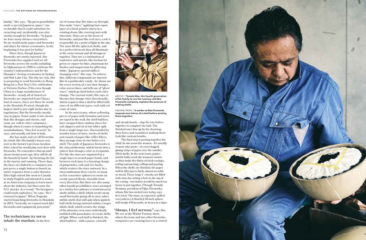 A man in a blue shirt holding two round firework shells with white wrapping and black and yellow markings, seated indoors with a plain wall behind him.