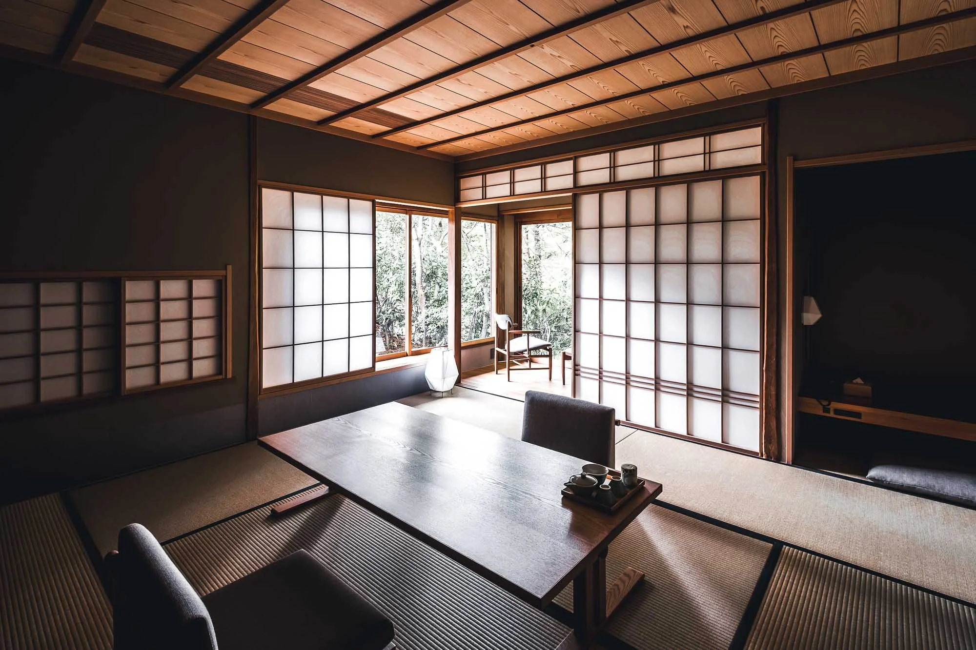 Interior of a Kyoto hotel room in traditional Japanese style, featuring shoji paper sliding doors, tatami mat flooring, and a low wooden table with dishes, illuminated by natural light from outside.