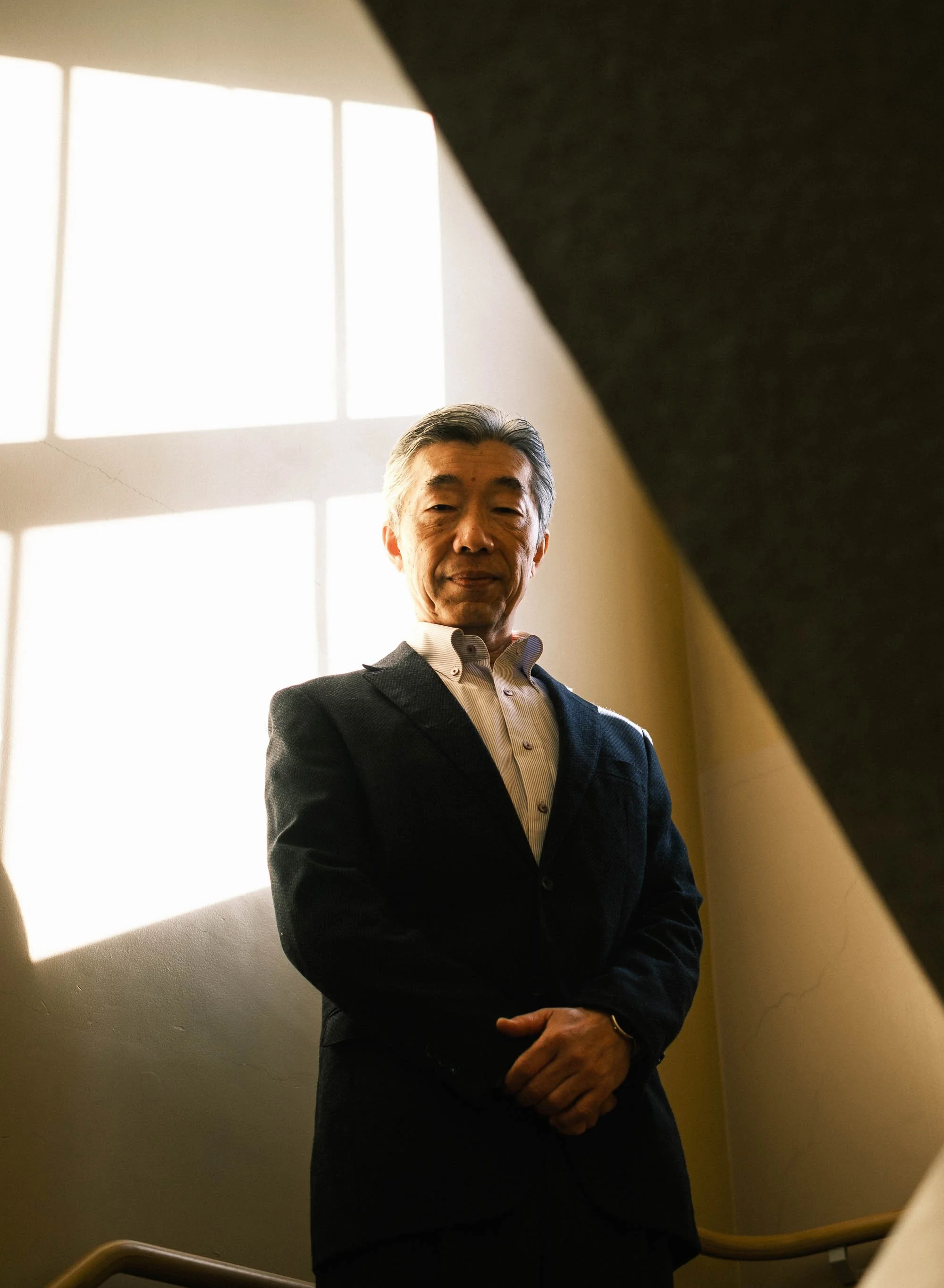 An editorial portrait of a Japanese Christian pastor in a dark suit and white shirt standing near a staircase against a yellow wall, illuminated by sunlight coming through a window.
