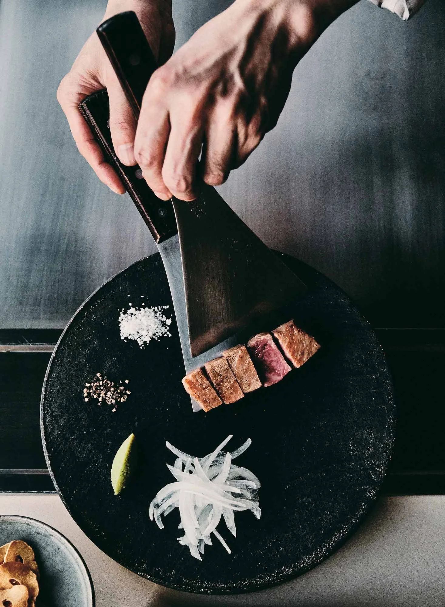 A chef serving grilled steak on a black stone plate with onion slices and salt and pepper on the side.
