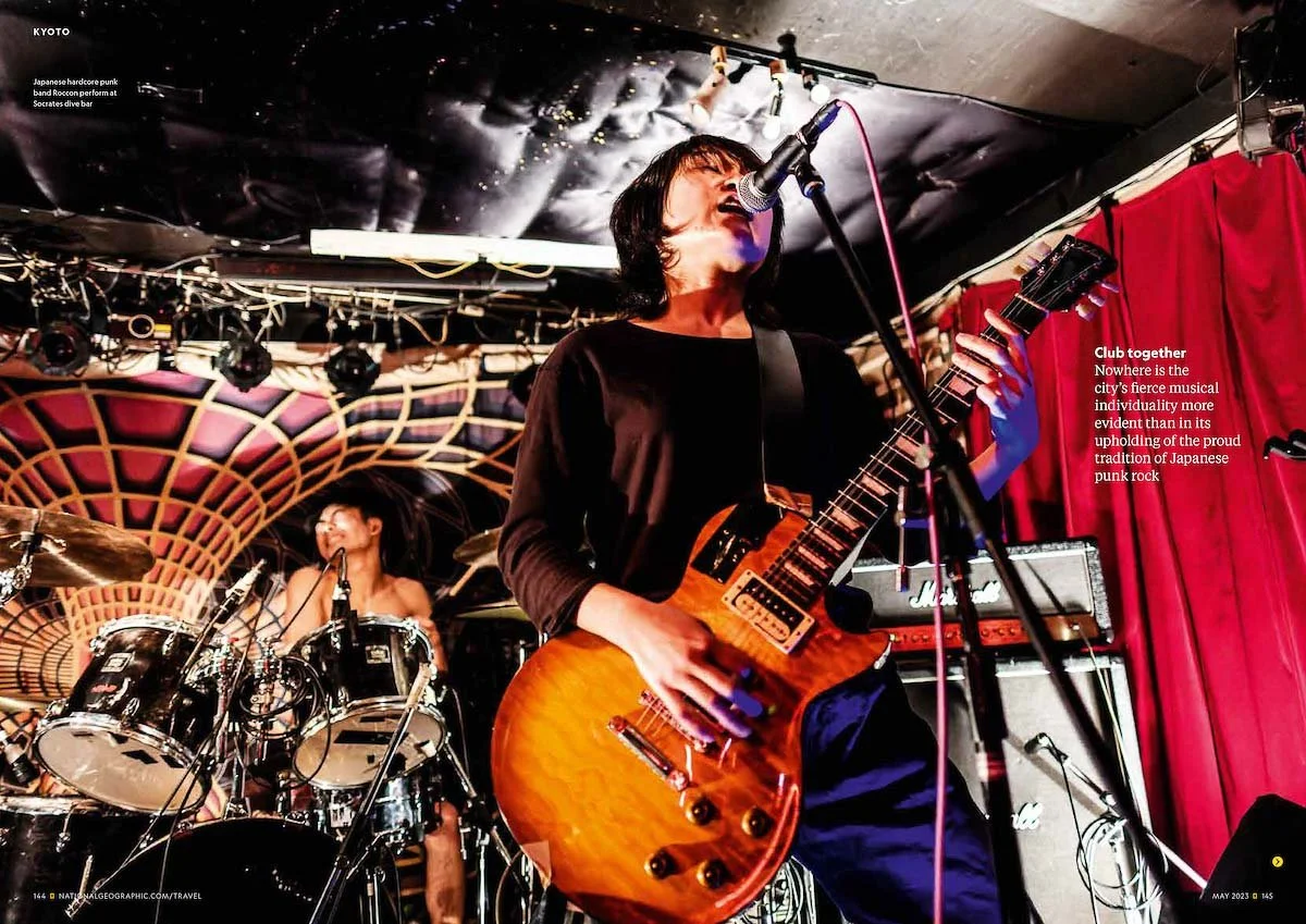 A musician with black hair singing into a microphone and playing an orange electric guitar on stage with a drummer in the background. The stage has acoustic band performance elements and a red curtain to the right.