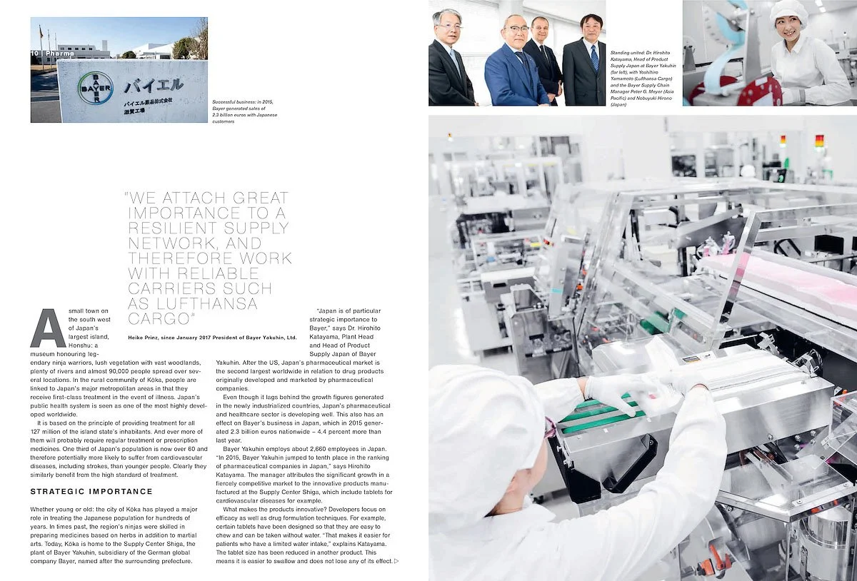 A group of five professionals in suits standing in a white, modern manufacturing or research facility, with one person wearing a protective hair cover, working on machinery.