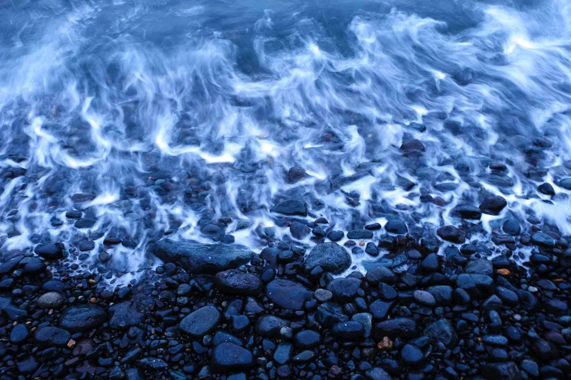 Waves washing over a rocky shoreline with small and large dark stones.