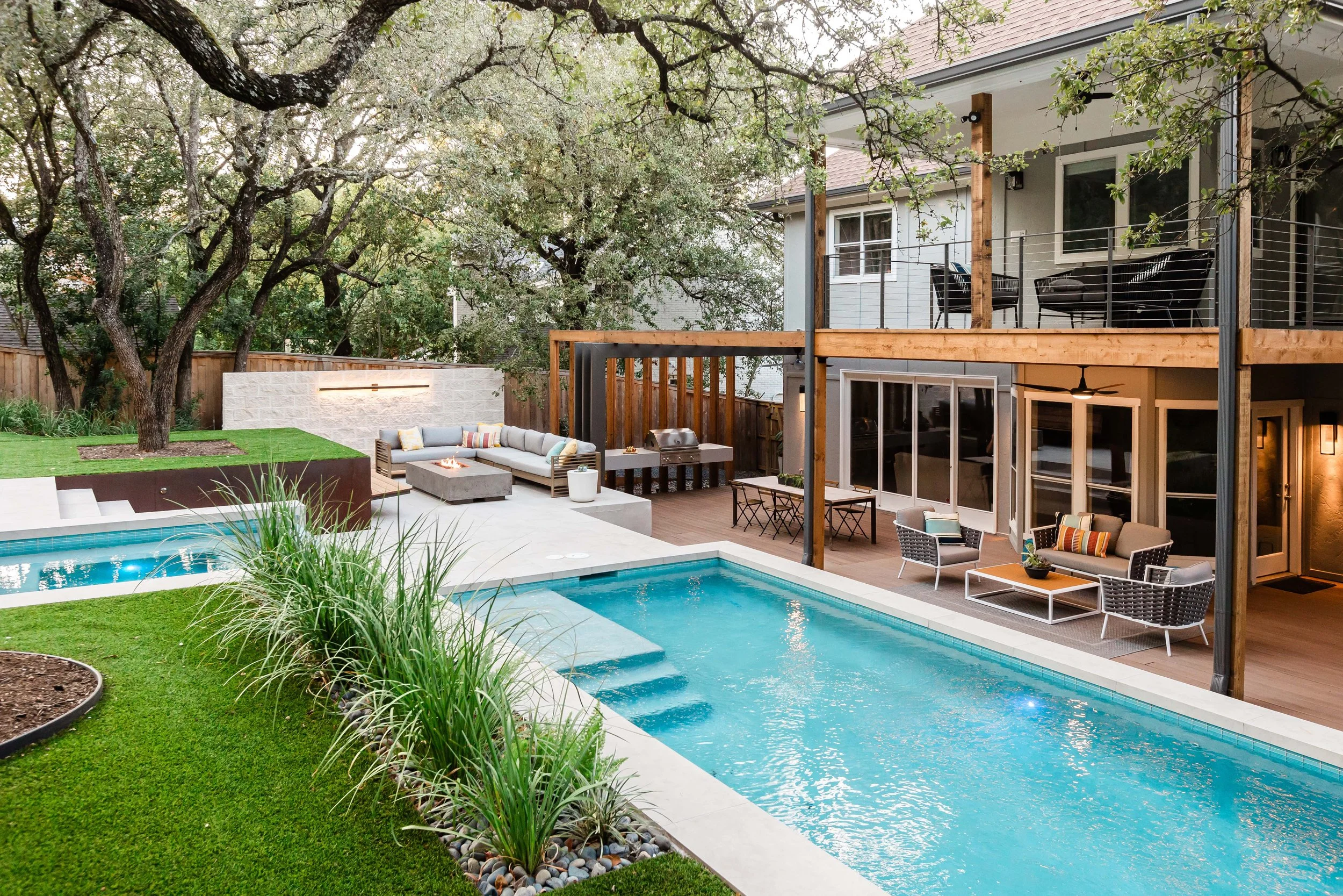 Long view of a modern pool and spa with outdoor lounge seating, framed by lush landscaping and a two-story home with a covered patio.
