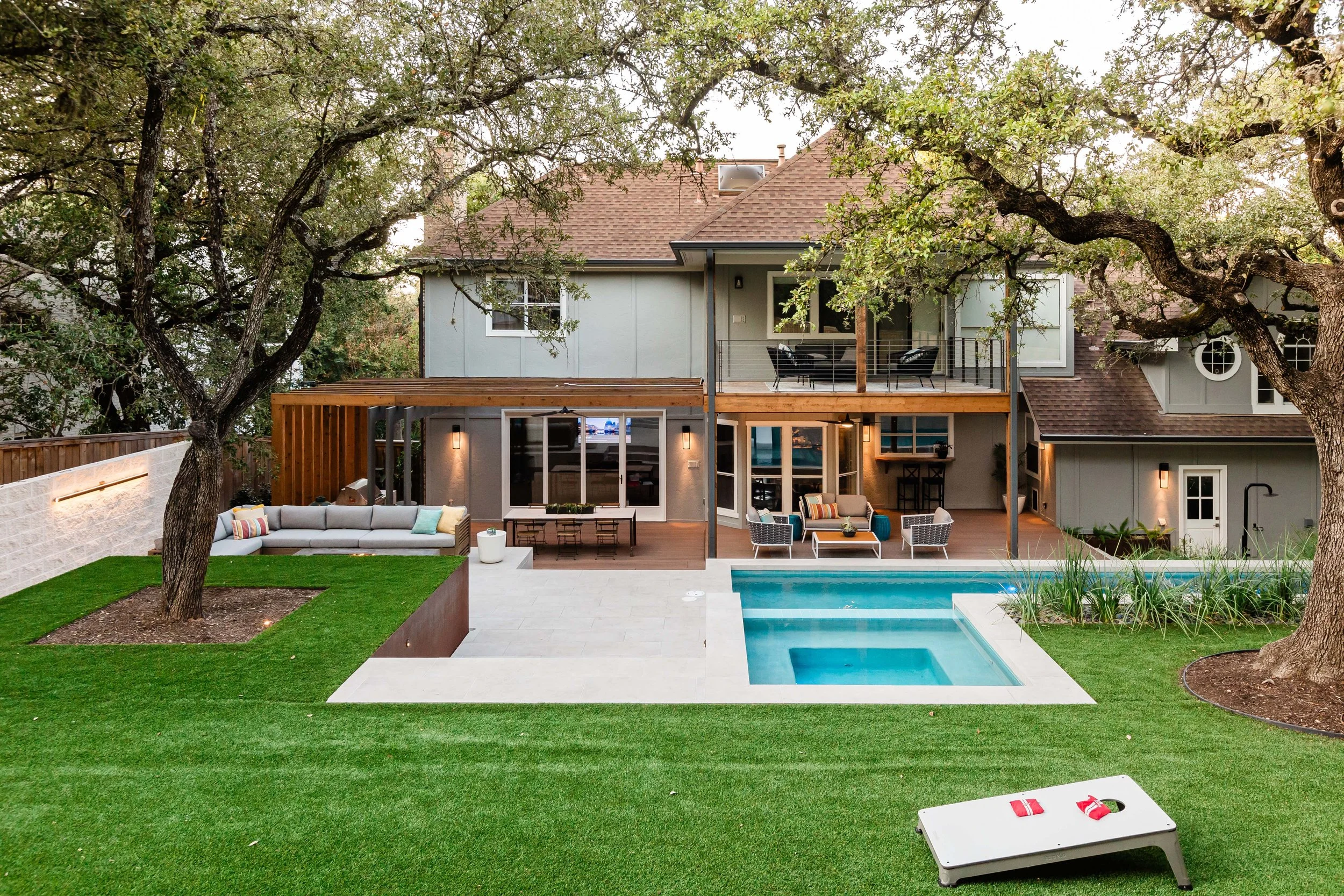 Modern backyard with a rectangular pool and hot tub, surrounded by a wood deck and outdoor living spaces beneath mature oak trees