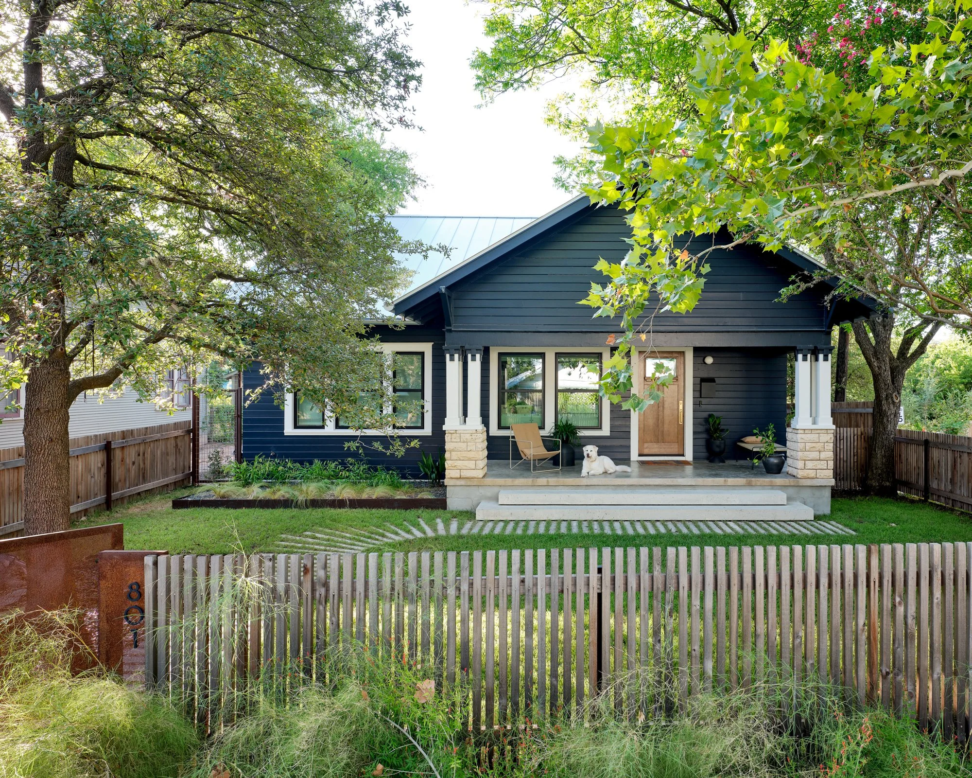 Navy-painted Craftsman home with light stone porch columns, vertical slat fence, linear-pattern stone walkway, and manicured front lawn