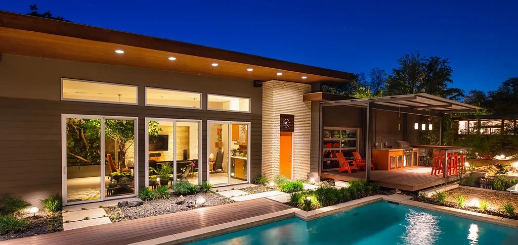 Modern house with large glass doors, lit patio, outdoor kitchen and dining area, and swimming pool at night.