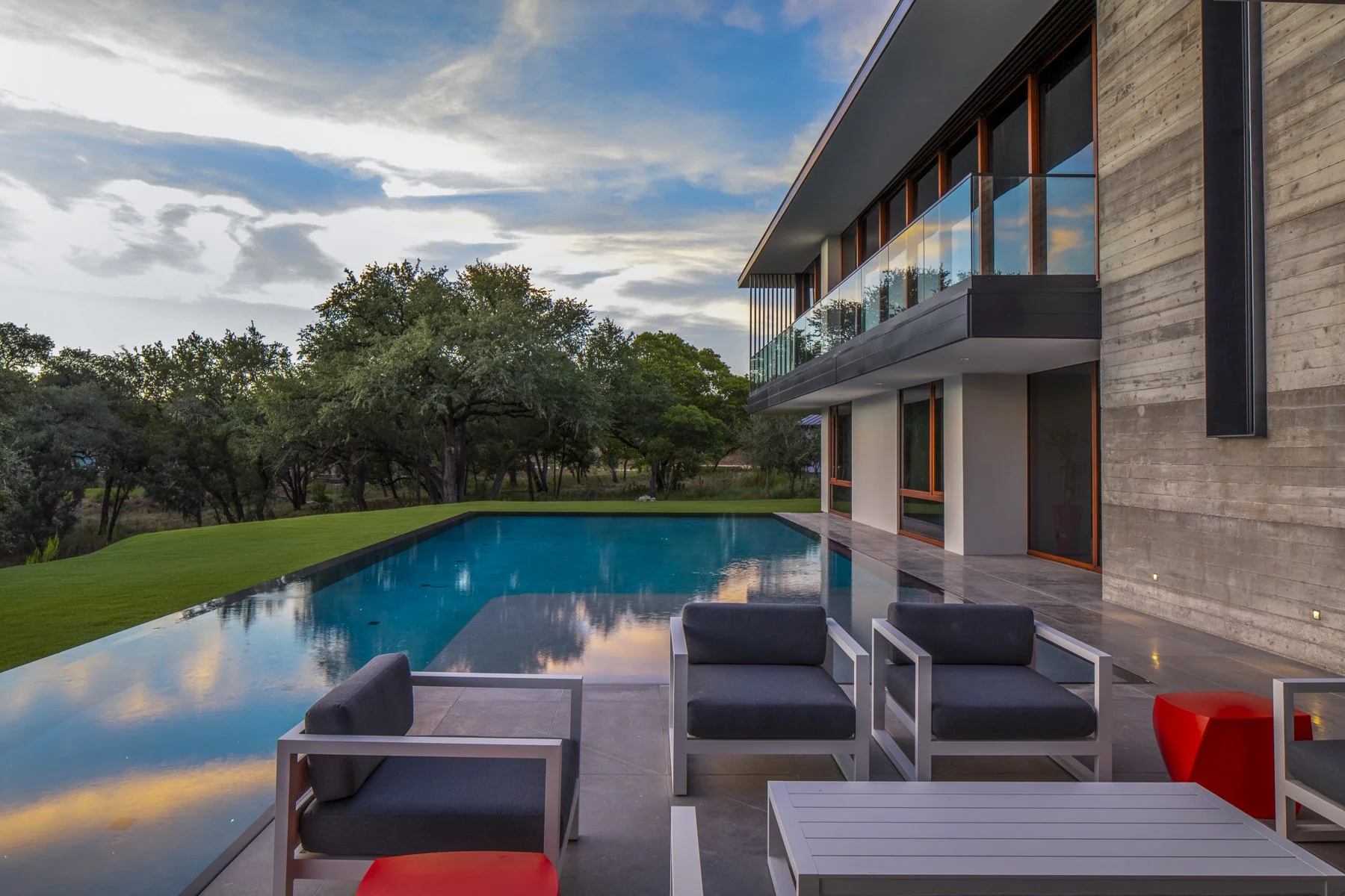 Sunset reflections ripple across a serene infinity-edge pool beside a modern home clad in board-formed concrete and glass, with sleek outdoor lounge seating overlooking lush Hill Country