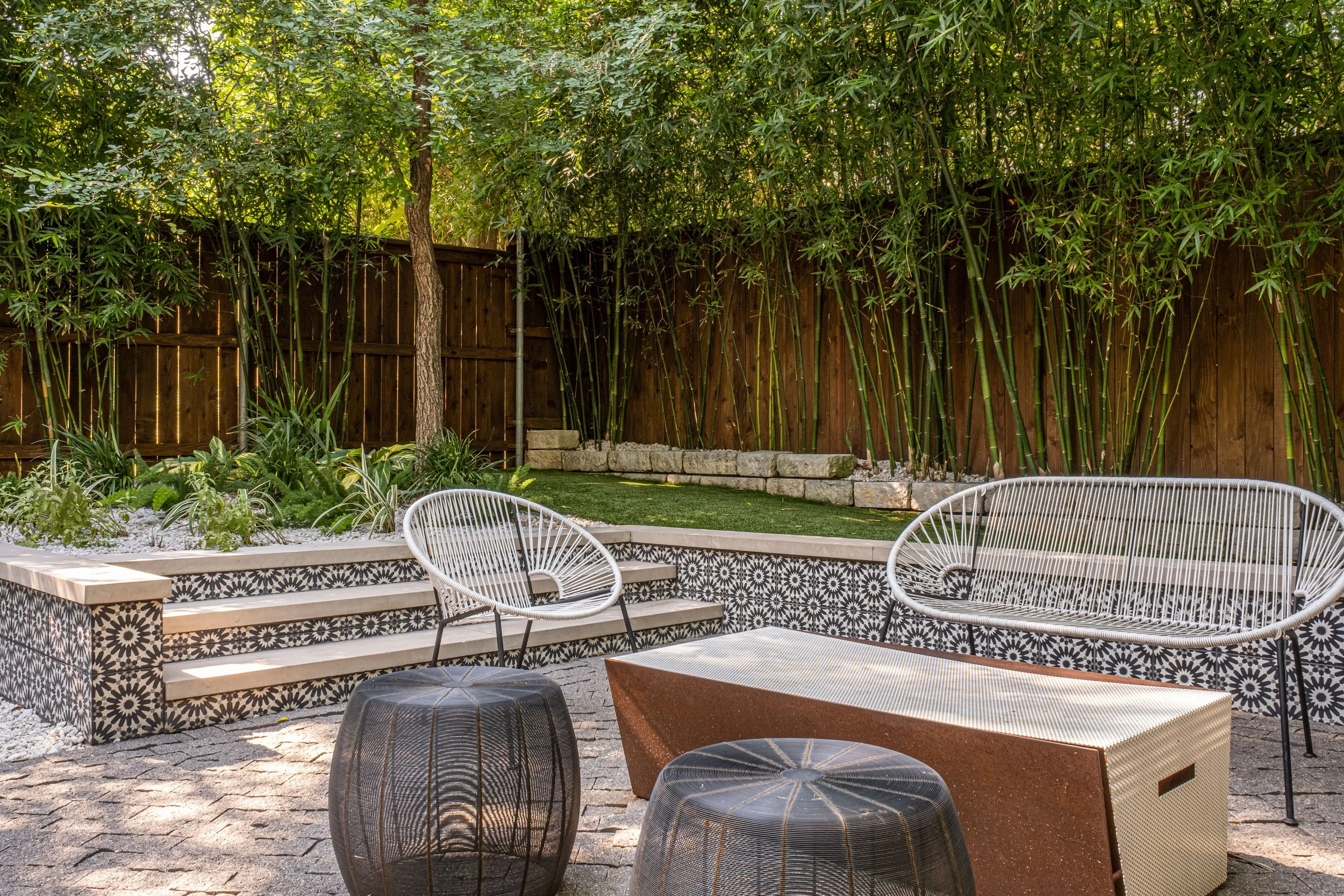 Tranquil seating nook in a bamboo-lined courtyard, featuring patterned tile retaining walls, modern wire-frame chairs, and a rusted steel fire pit centerpiece—layered with natural textures and softened by dappled sunlight