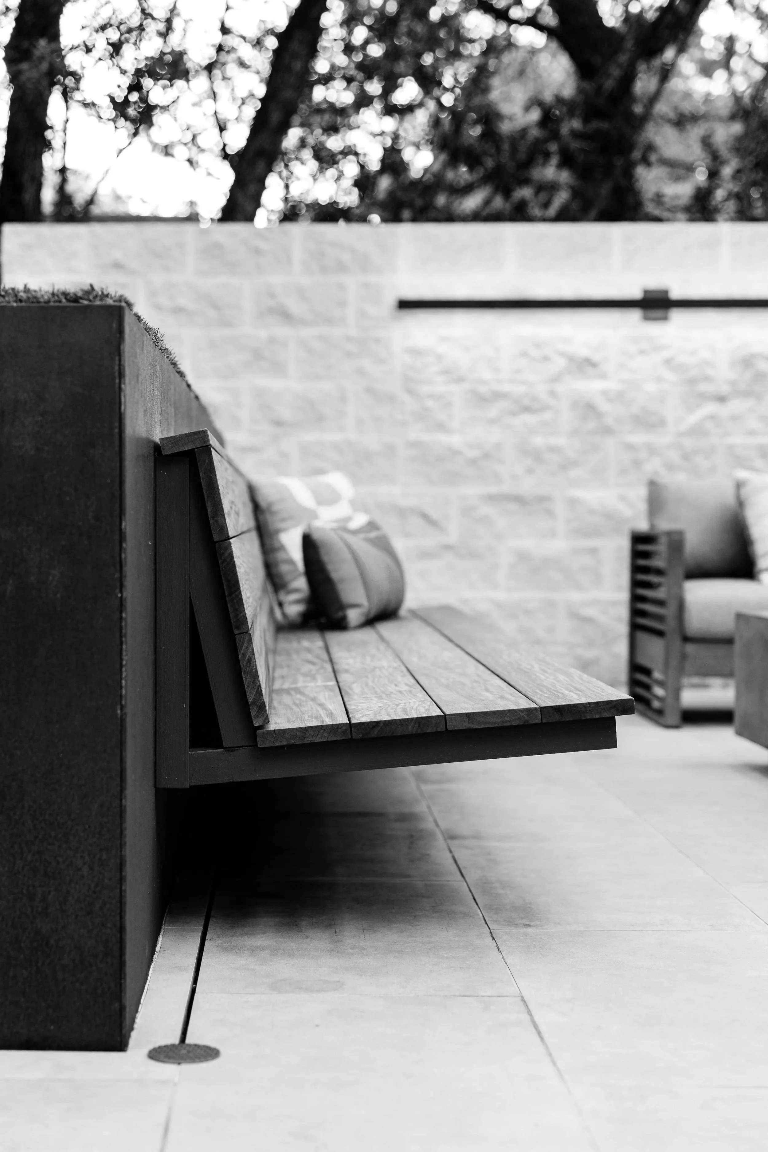 Black and white photo of a wooden outdoor bench with cushions on it, placed against a short steel wall. Privacy wall with modern light fixture.