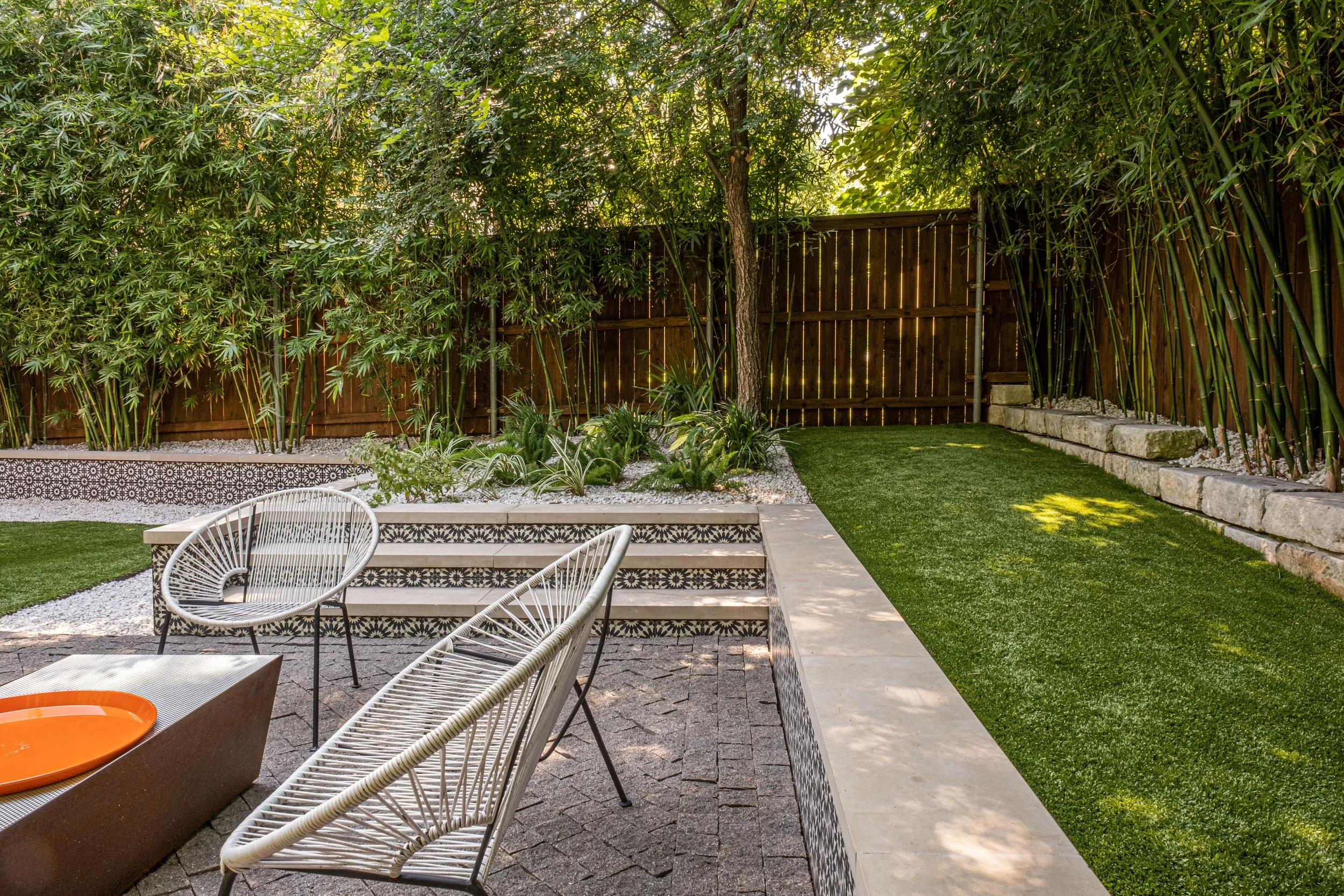 Modern outdoor lounge with white woven chairs, a low fire pit, and decorative tile planters, adjacent to an artificial turf lawn bordered by bamboo and native greenery
