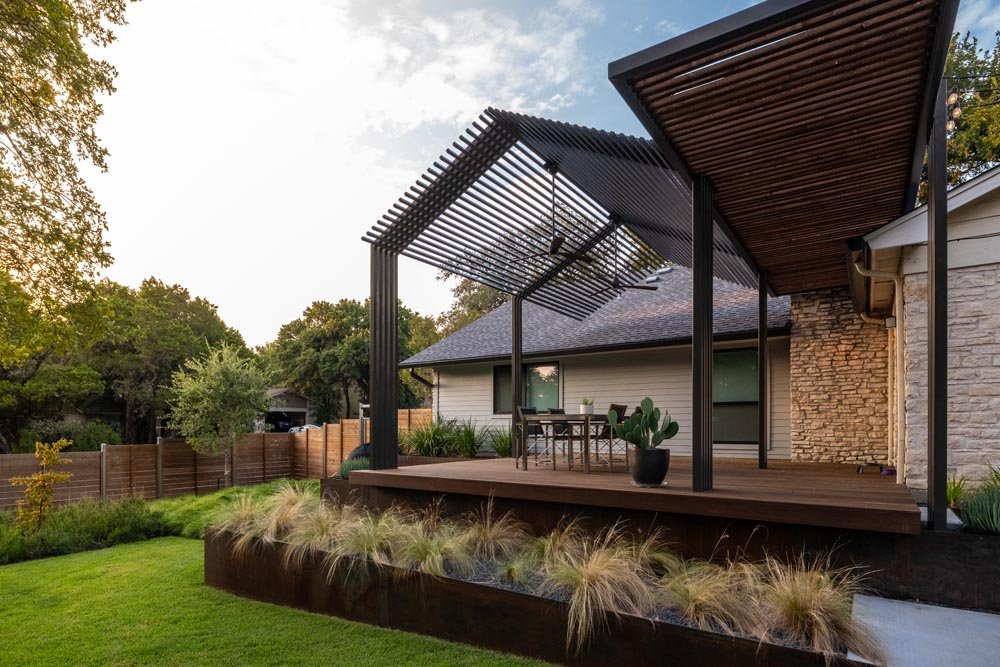 Modern backyard deck with geometric steel pergola structure, elevated above steel planters filled with ornamental grasses, all set against a backdrop of warm stone siding and lush green lawn